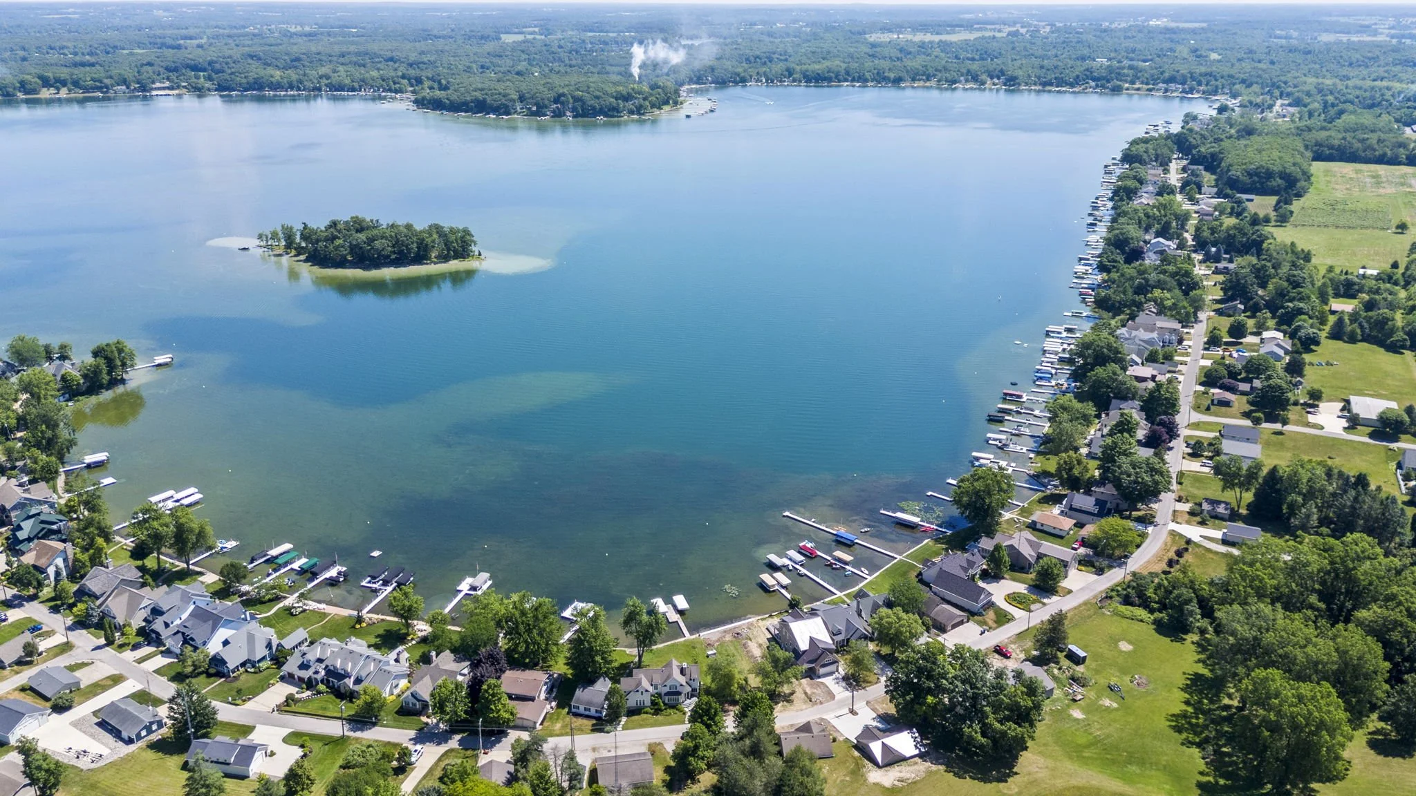Aerial view of a lakeside residential area with houses, docks, and boats along the shoreline, surrounded by lush green trees and water.