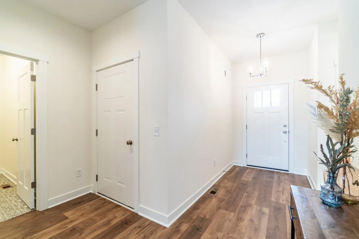 Empty white-walled entryway with front door, side walls, wooden flooring, and a vase with dried flowers on a wooden console table.