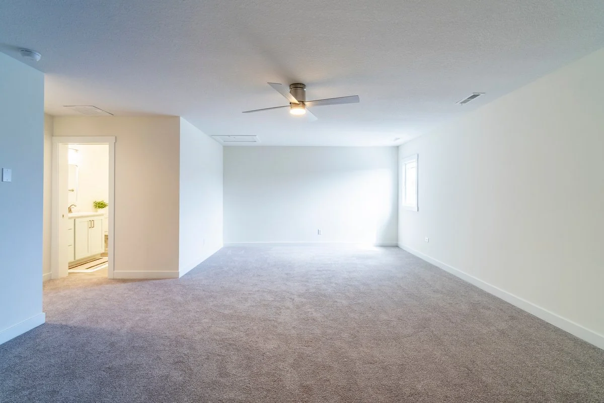 Empty living room with beige carpeting, white walls, a ceiling fan with a light, and a window allowing natural light. There's a small bathroom with a sink and cabinets visible through a doorway.
