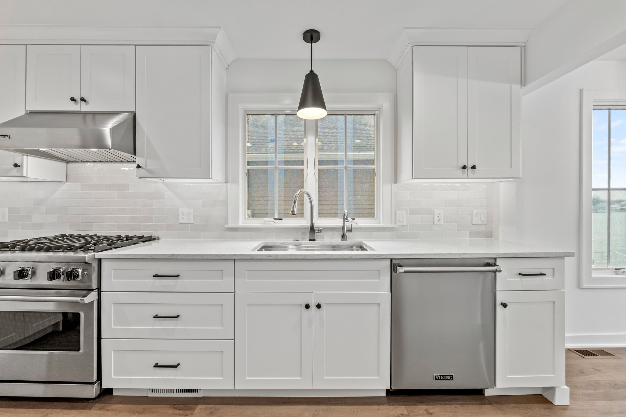 Modern white kitchen with stainless steel appliances, including an oven, dishwasher, and range hood, with a window above the sink letting in natural light.