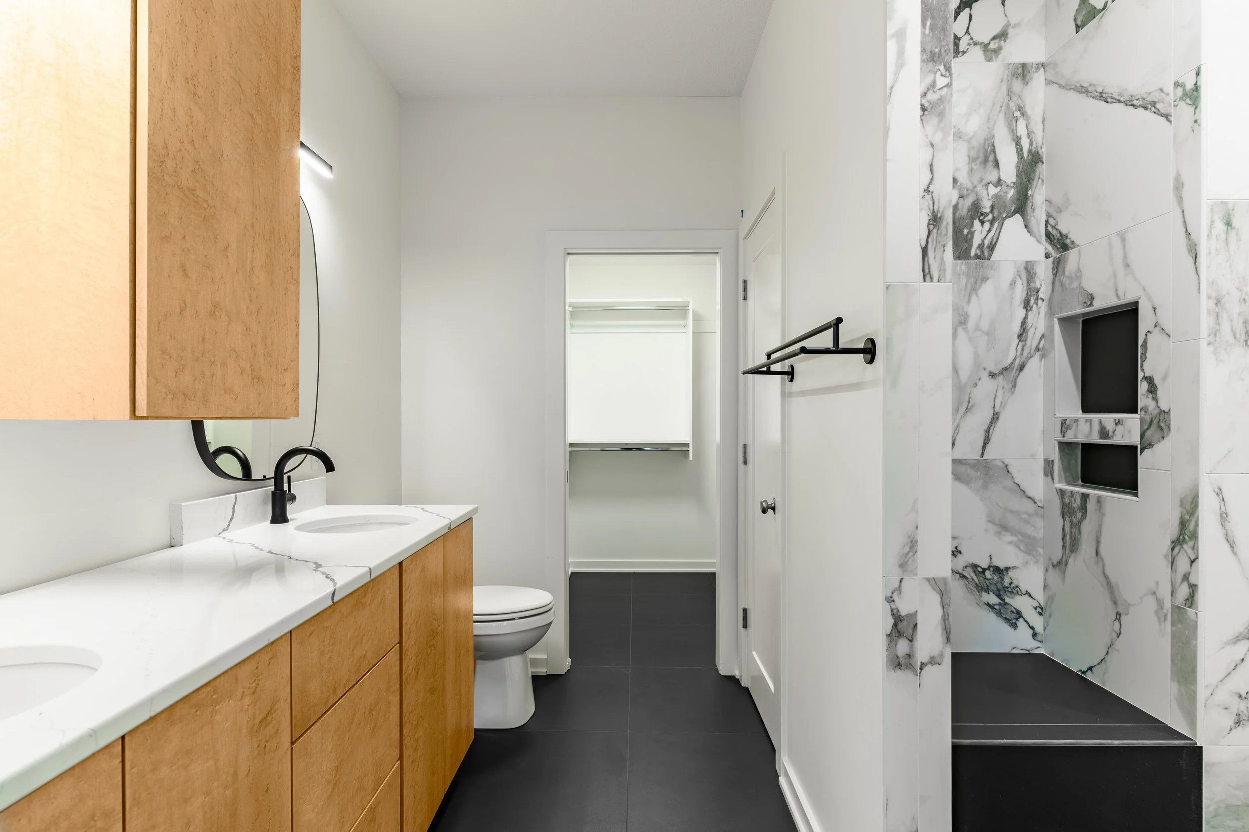 Modern bathroom with double vanity, black faucet, and large mirror, toilet, black towel rack, walk-in shower with marble walls and built-in niches, black floor tiles, and white walls.