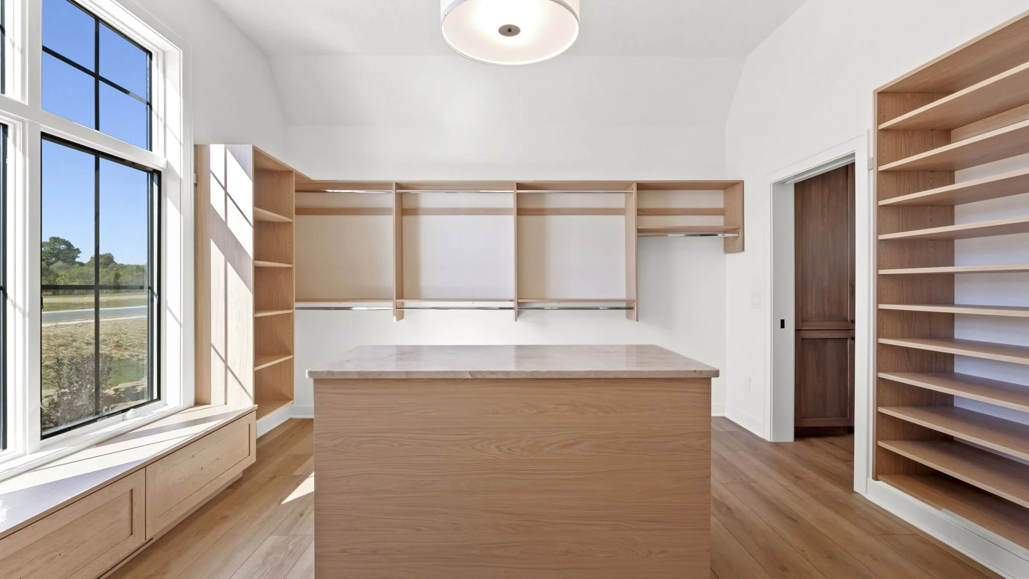 An empty walk-in closet with wooden shelves, a large window on the left, and a wooden island in the center. The closet has white walls and a hardwood floor.