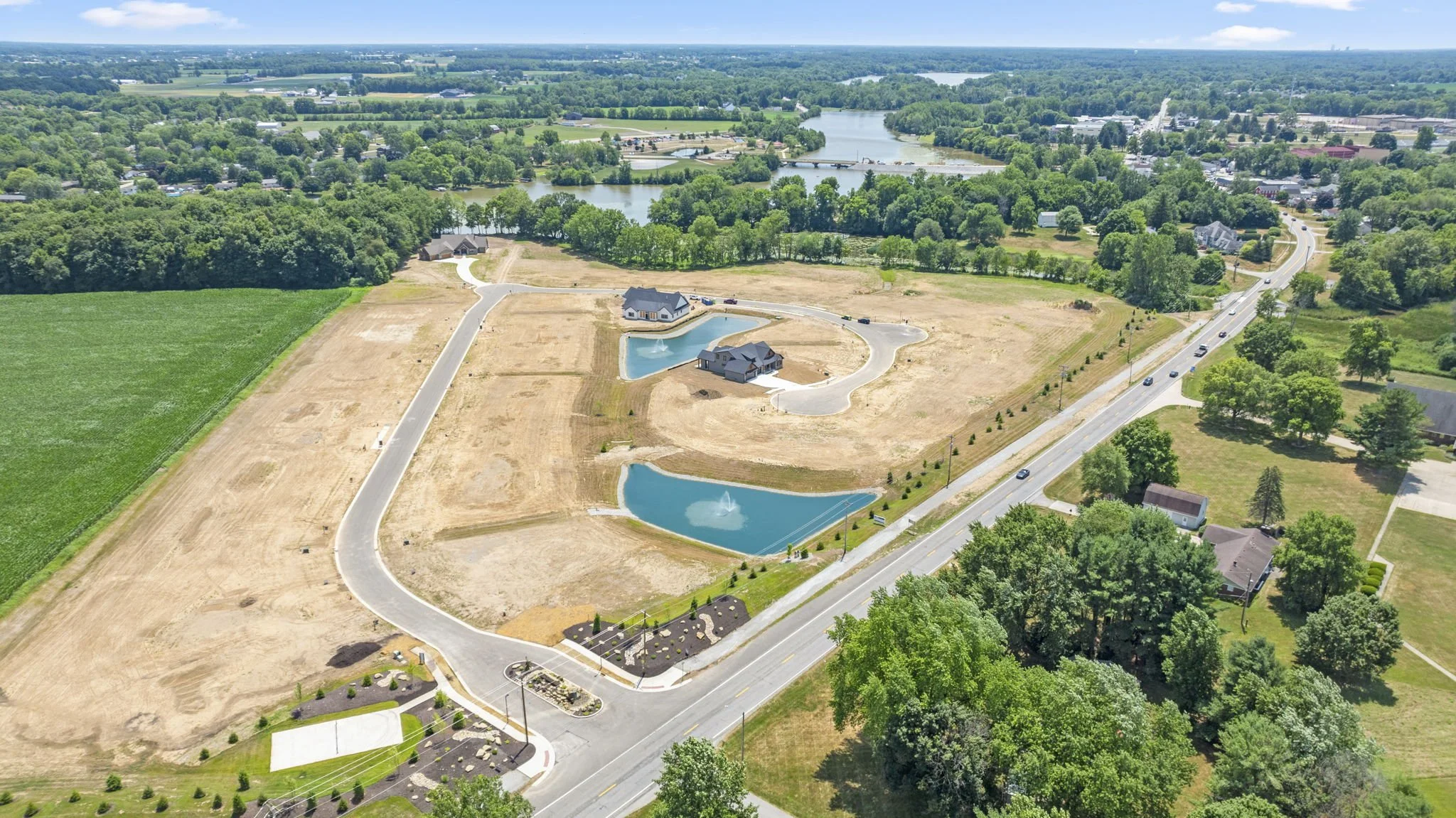 Aerial view of a suburban area with two small lakes, fountains, houses, roads, and green trees, with a river in the background.