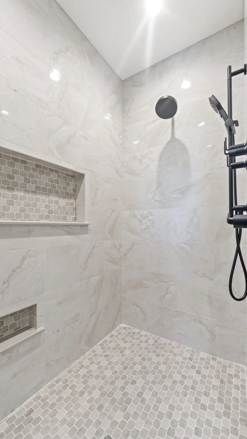The image shows a modern, tiled walk-in shower with beige marble tiles on the walls and small hexagonal tiles on the floor. There are built-in niches on the wall with smaller hexagonal tiles, and a black showerhead with a handheld shower attachment i