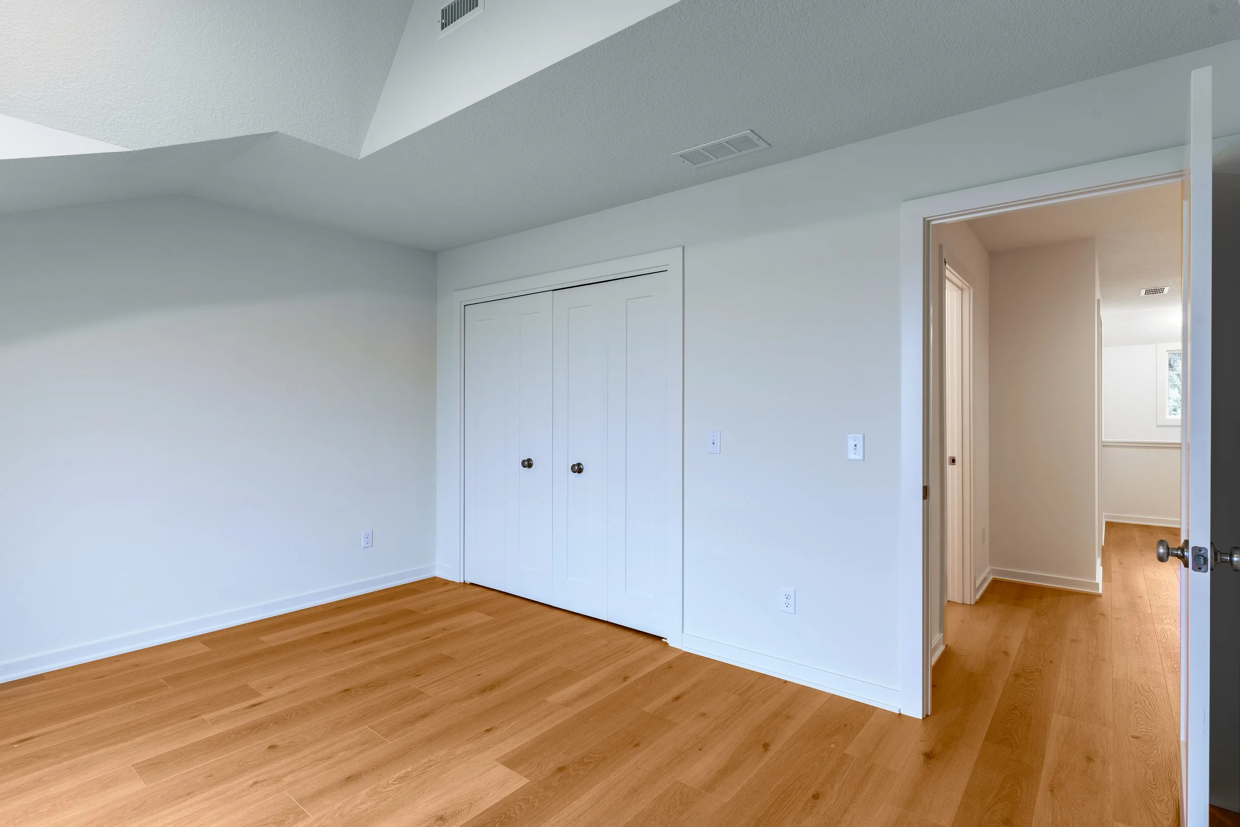 Empty room with white walls, wooden floors, a closet with two white doors, and an open doorway leading to a hallway with more wooden floors and a window.
