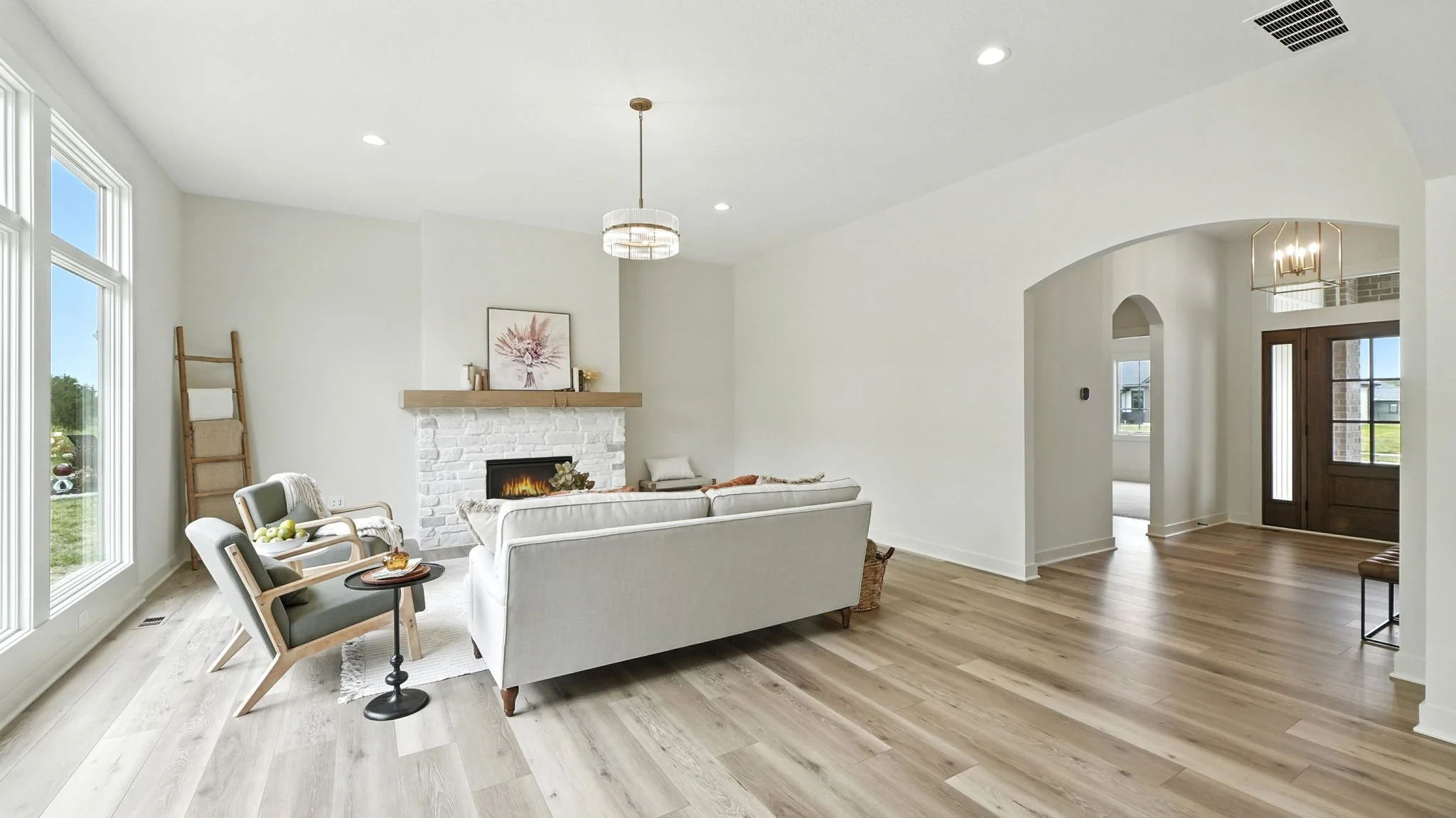 Living room with light wood flooring, white sofa, armchairs, white brick fireplace, and large windows with a view outside.