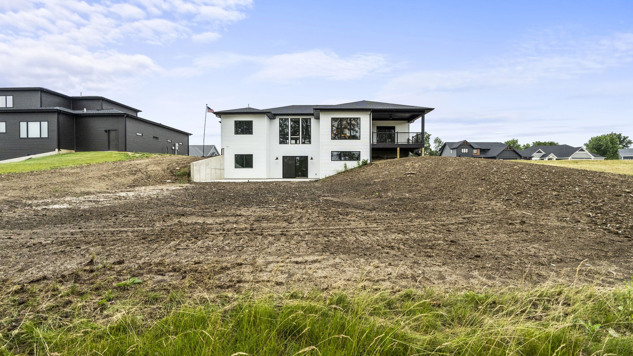 A large, newly built white house with a black roof and a balcony, situated behind a dirt field, with neighboring houses and a partly cloudy sky in the background.