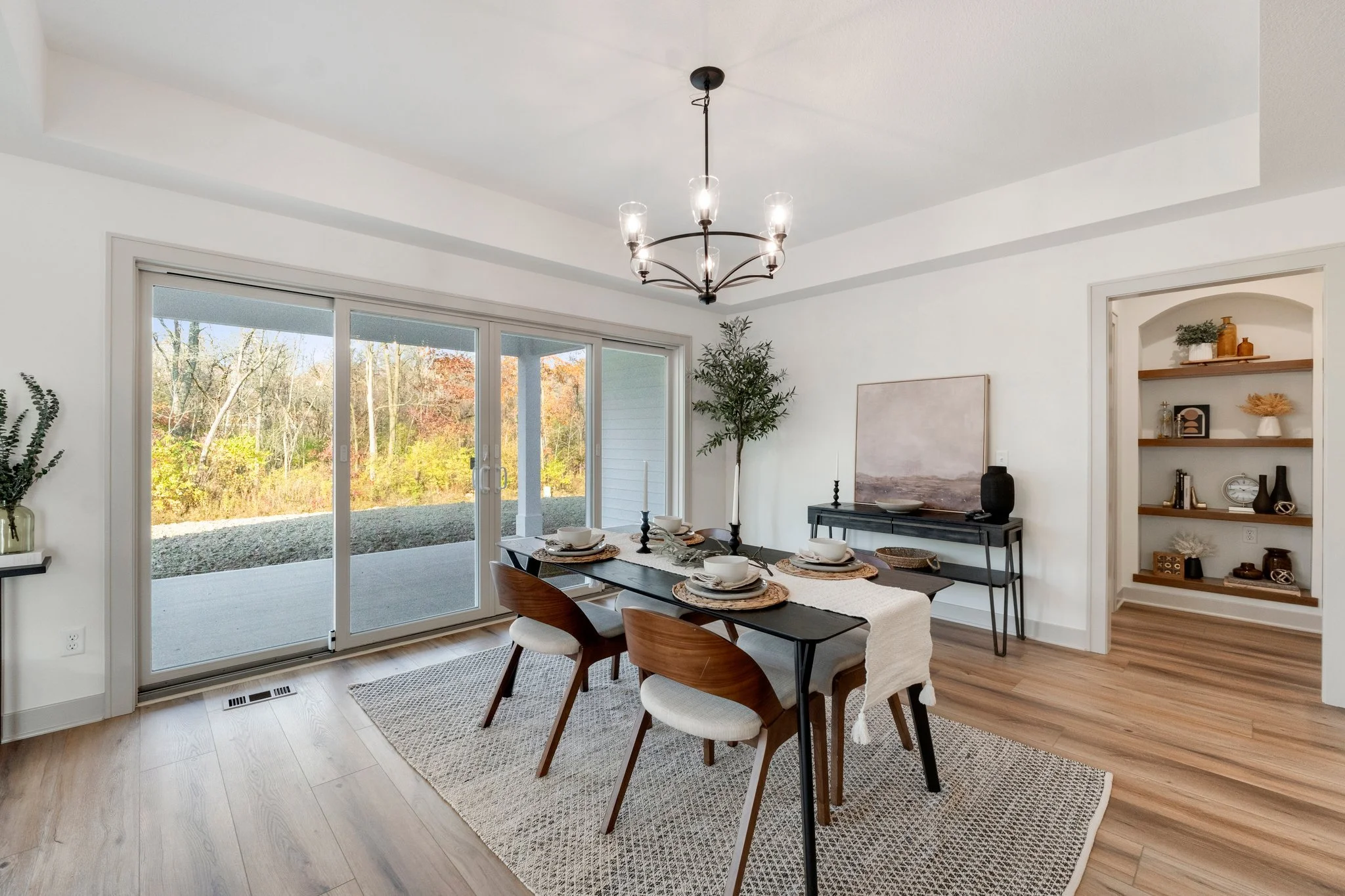 Dining room with a black table set with four place settings, wooden chairs, a modern chandelier, sliding glass door leading outside, a black sideboard with artwork, and built-in shelves with decorative items.