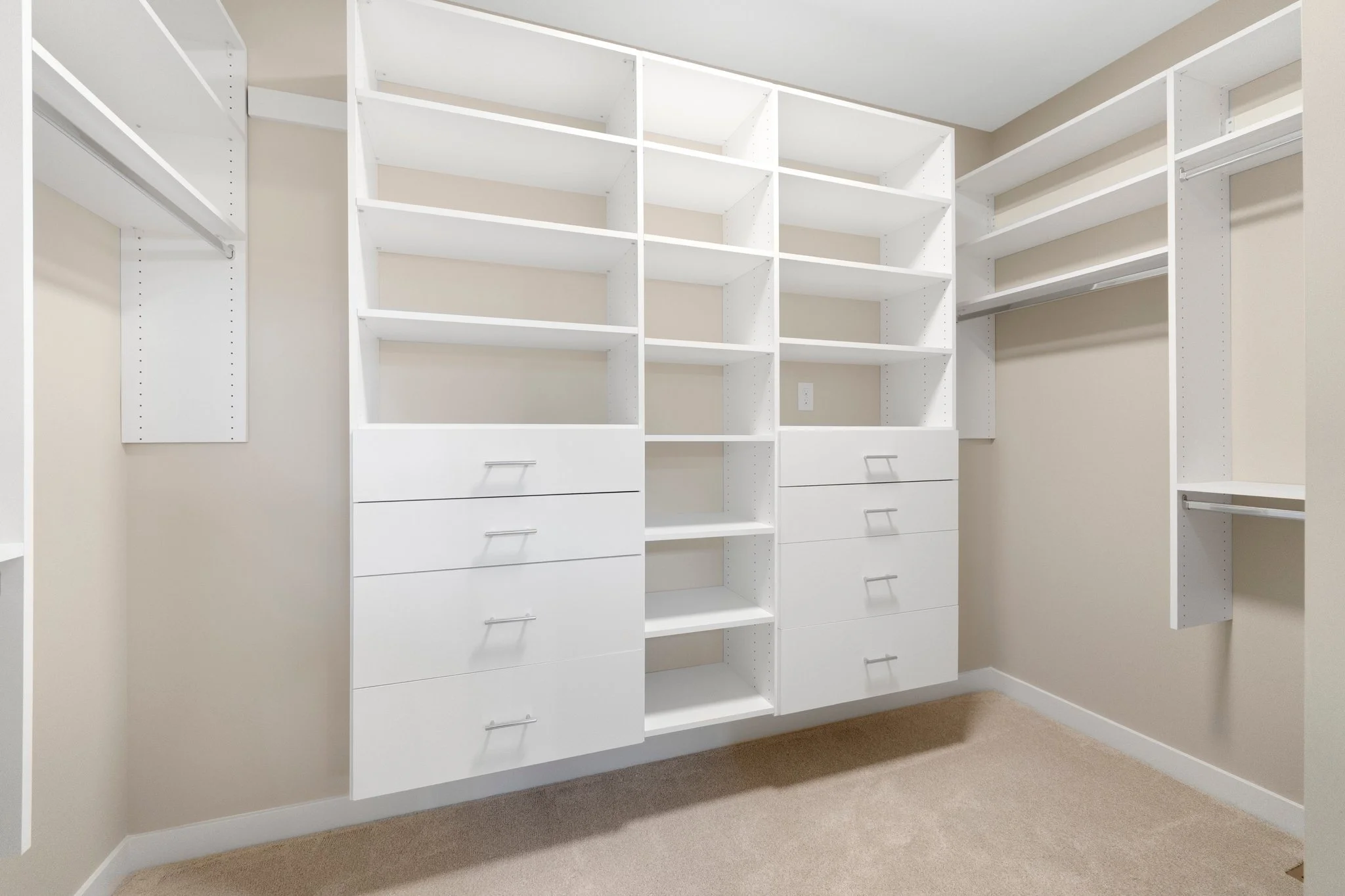Empty walk-in closet with white shelving units, drawers, and beige walls, carpeted floor.