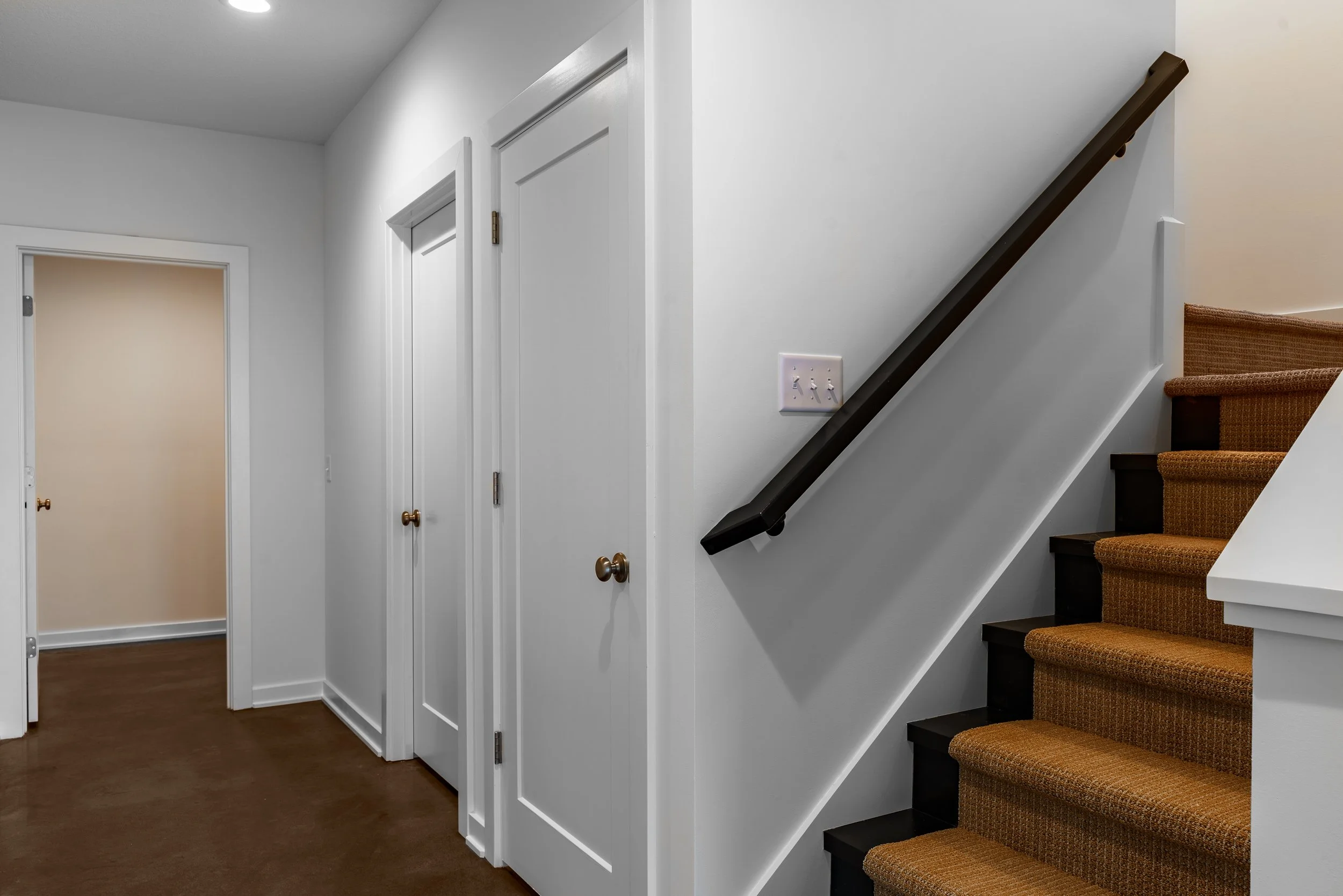 Interior view of a home with a staircase on the right, carpeted stairs, white walls, and closed doors on the left.