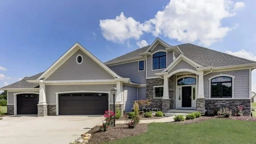 Large modern house with three garages, stone accents, and landscaped yard under a partly cloudy sky.