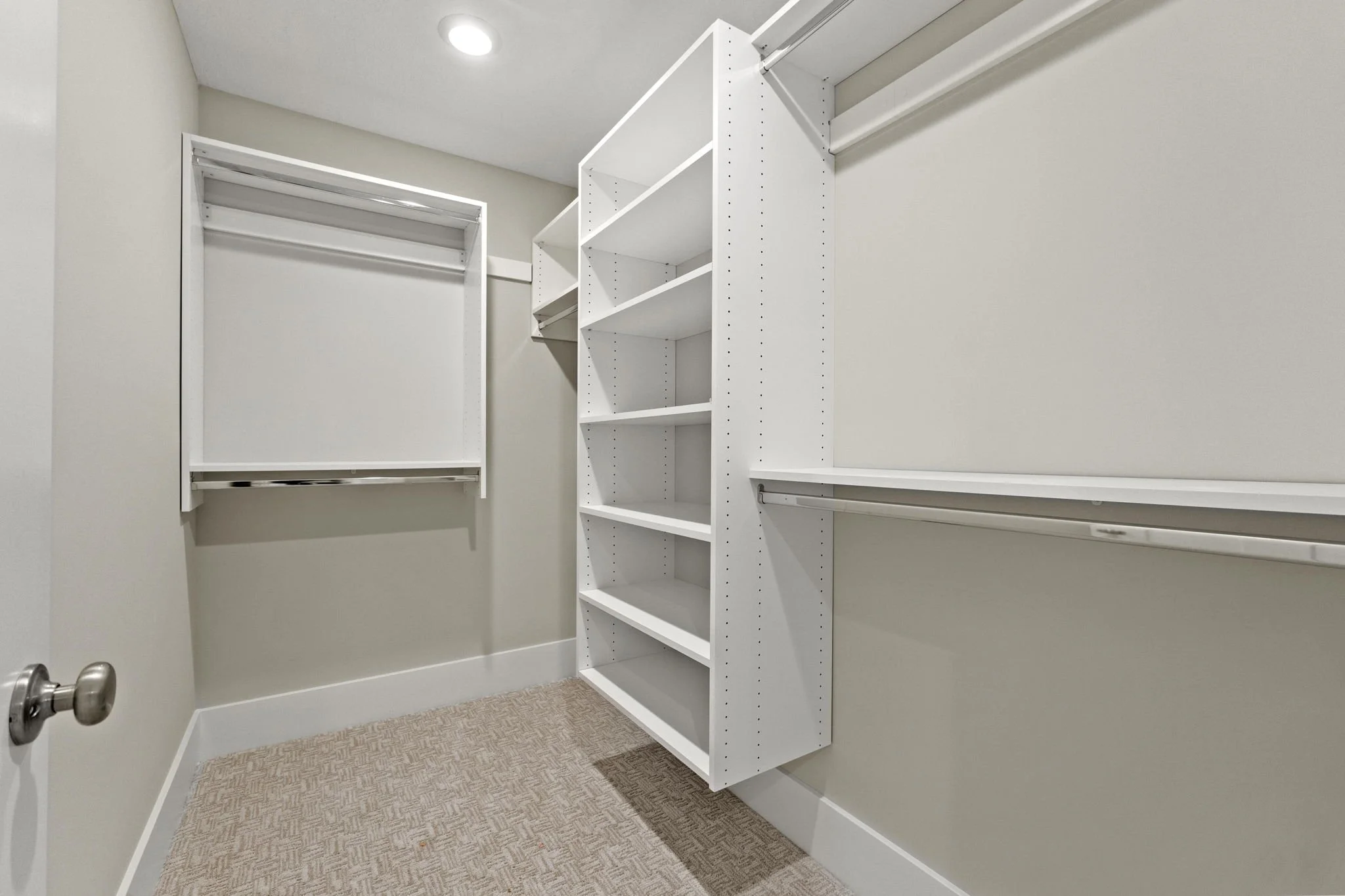 Empty walk-in closet with white built-in shelves and hanging rods, beige carpet, and neutral-colored walls.