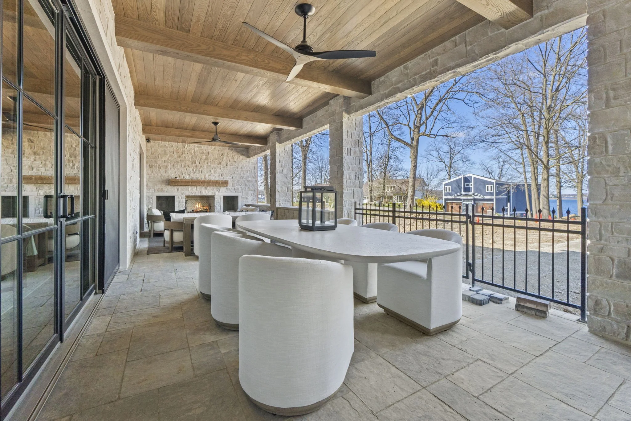 An outdoor patio with white chairs around a rectangular table, a lantern on the table, and a fireplace in the background. The patio has a wooden ceiling and stone flooring, with trees and a building visible outside.