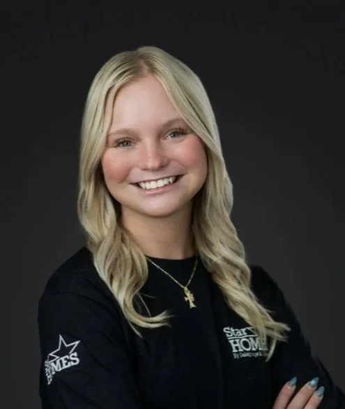A young woman with long blonde hair smiling, wearing a black shirt with logo and a gold necklace, standing against a dark background.