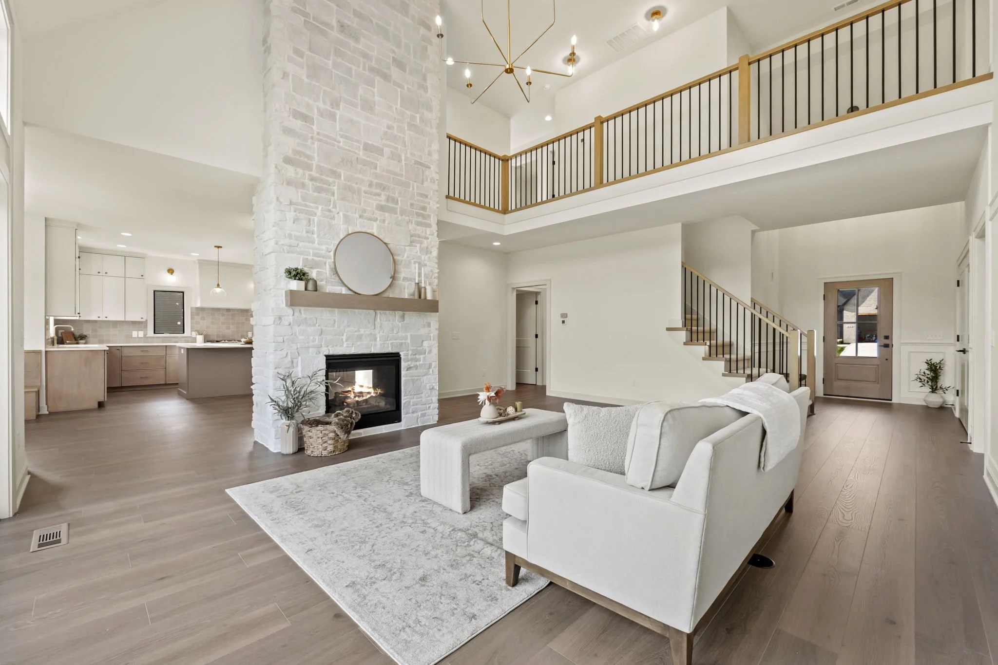 Open-concept living room with white walls, a white couch, a light-colored area rug, a fireplace with a white stone chimney, and a staircase with wooden steps and black metal railing leading to a second-floor loft. The room features modern decor and n