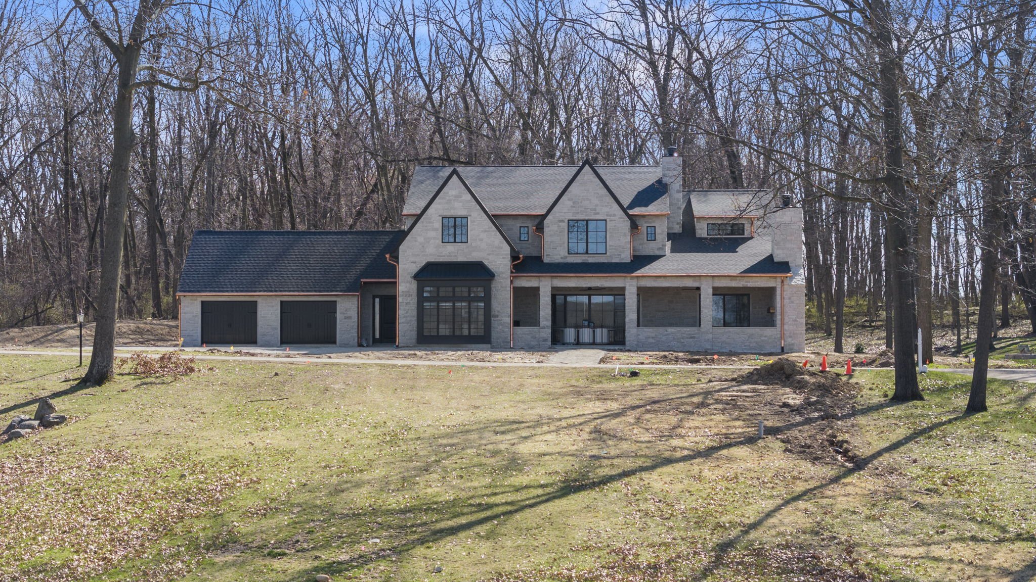A new two-story house under construction with a brick exterior, large windows, a two-car garage, and a covered porch, surrounded by trees and a lawn.