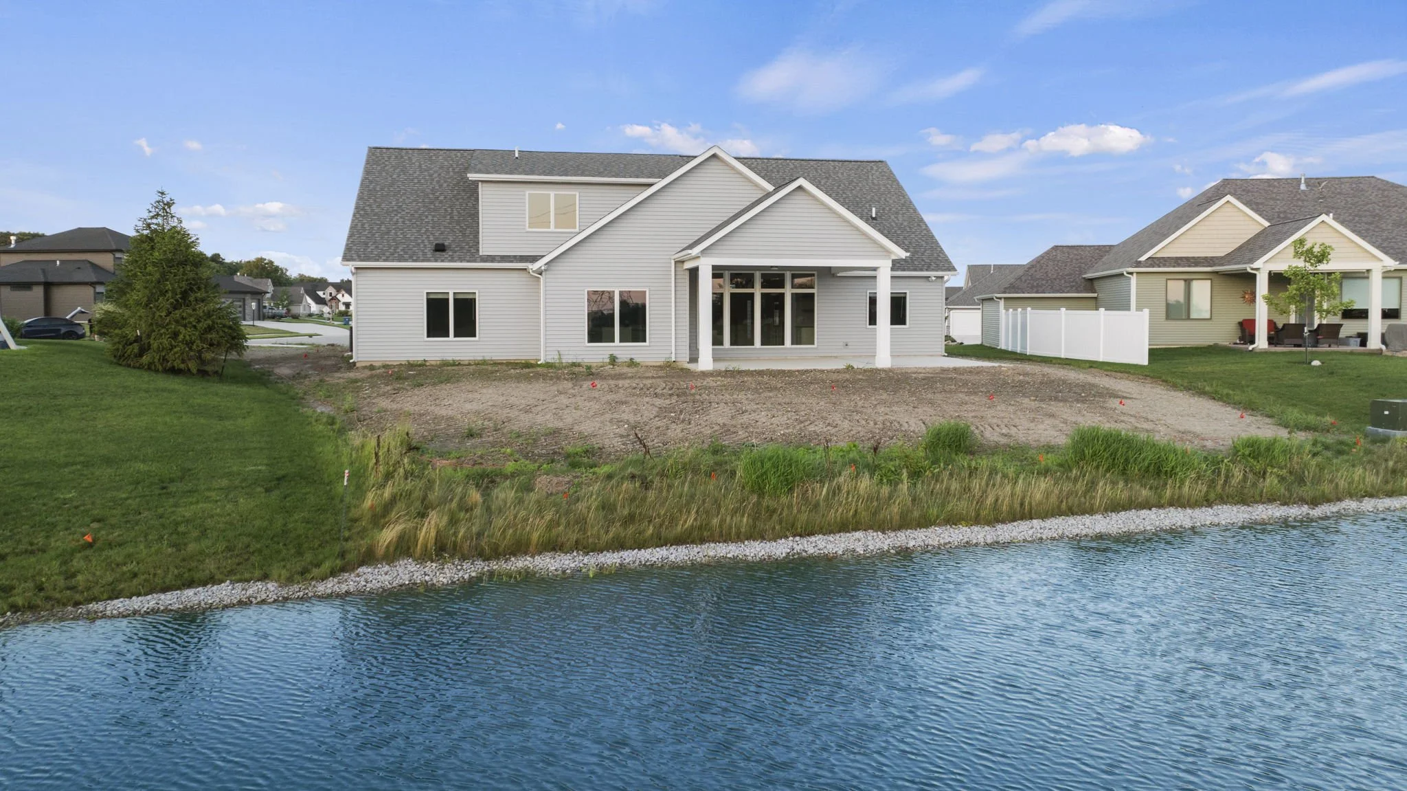 A white two-story house with a gable roof, situated on a grassy lot by a body of water under a partly cloudy sky.
