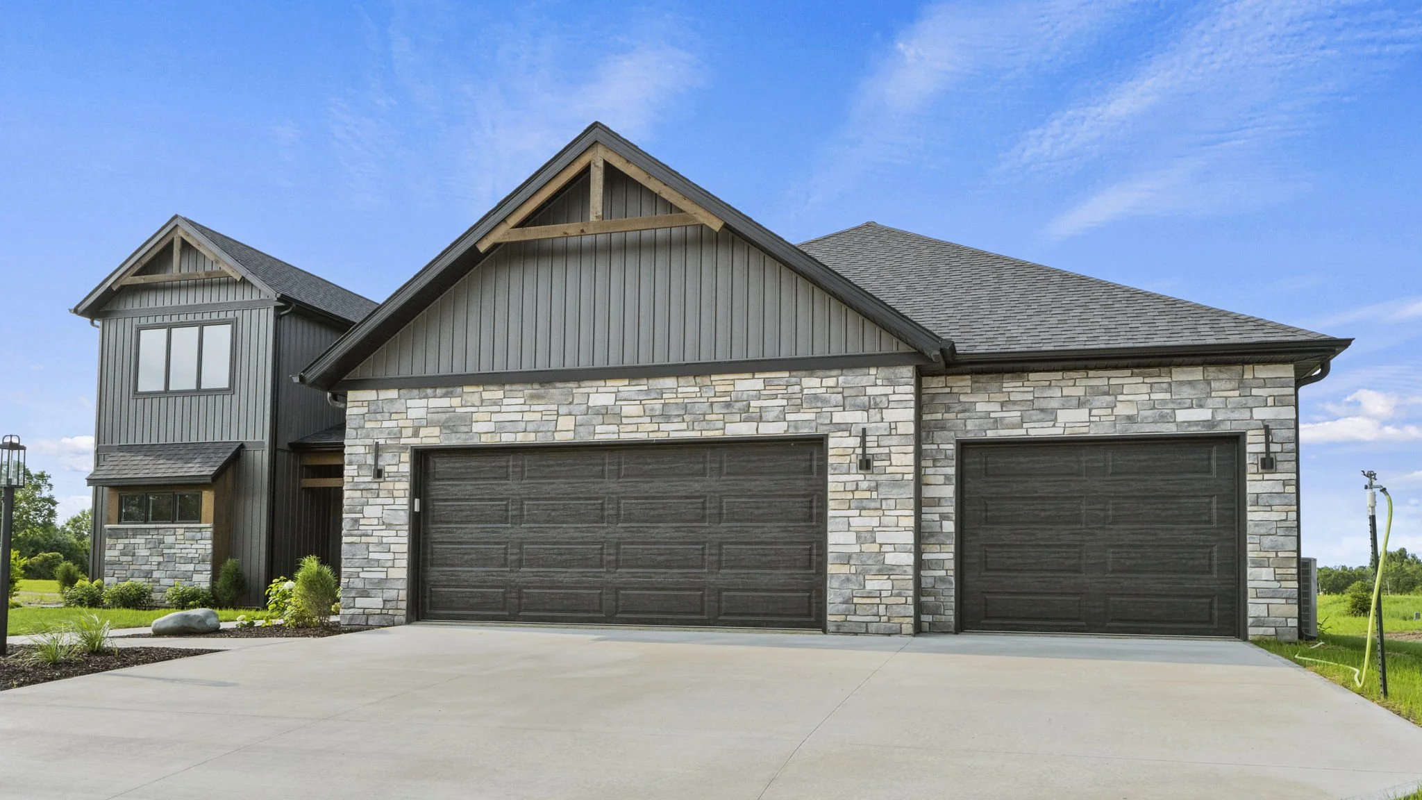 Modern house with two garage doors, stone facade, dark siding, and a pitched roof, under a blue sky.