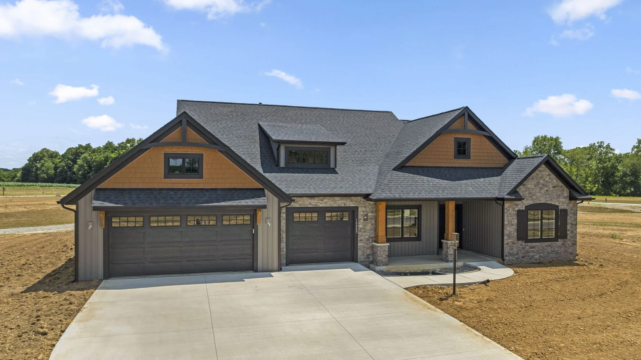 Newly built modern house with black garage doors, gray siding, stone accents, wooden front porch columns, and a concrete driveway in a rural setting with clear skies.