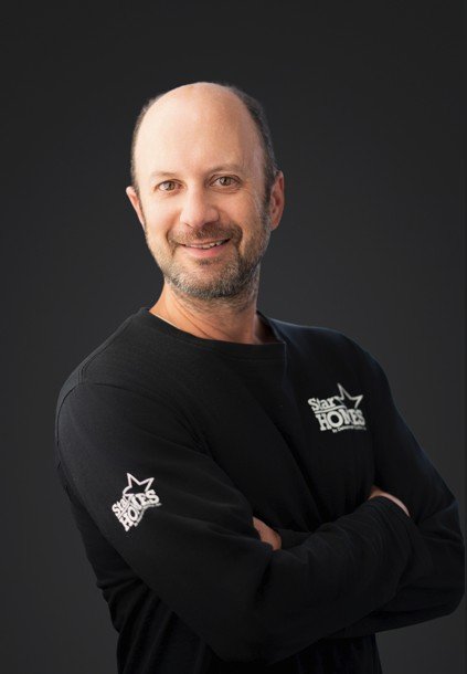 A middle-aged man with a beard and bald head smiling, wearing a black sweatshirt with 'Stars Homicide' logos, standing with arms crossed against a dark background.