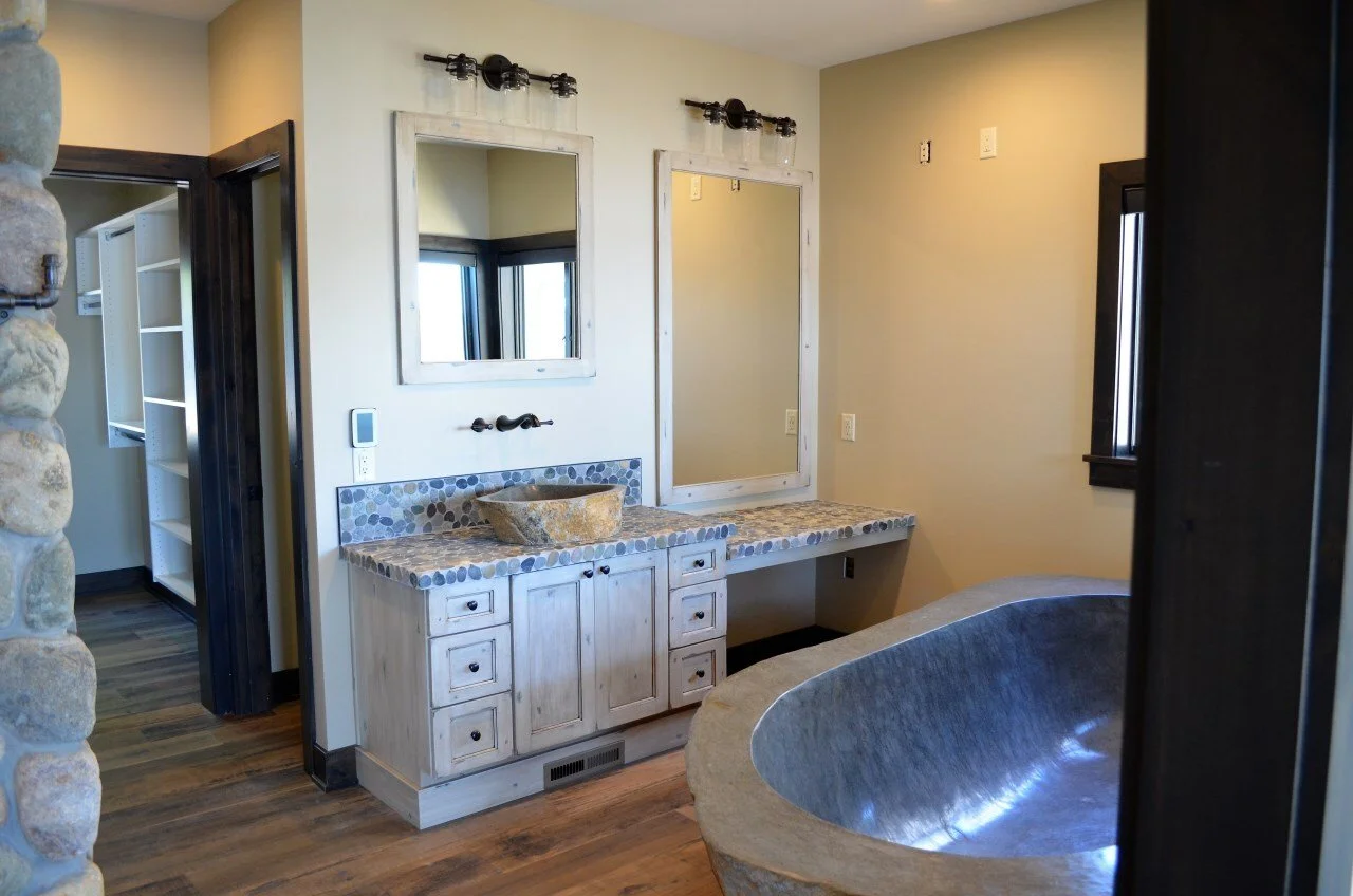 Rustic bathroom with double vanity, stone countertops, mirrors, and a large stone bathtub.