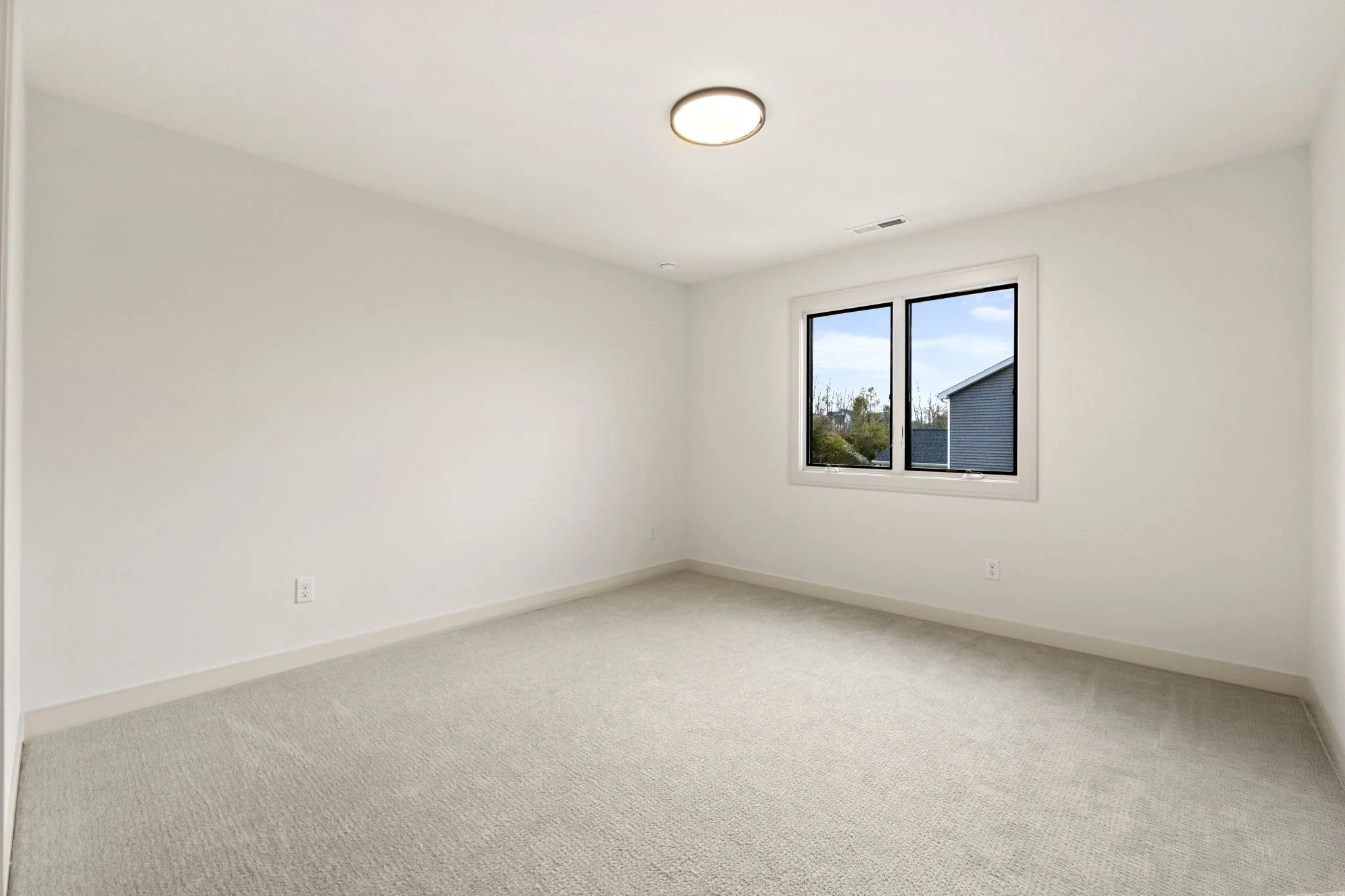 Empty room with white walls, beige carpet, a window showing neighboring house and sky, and ceiling light fixture.