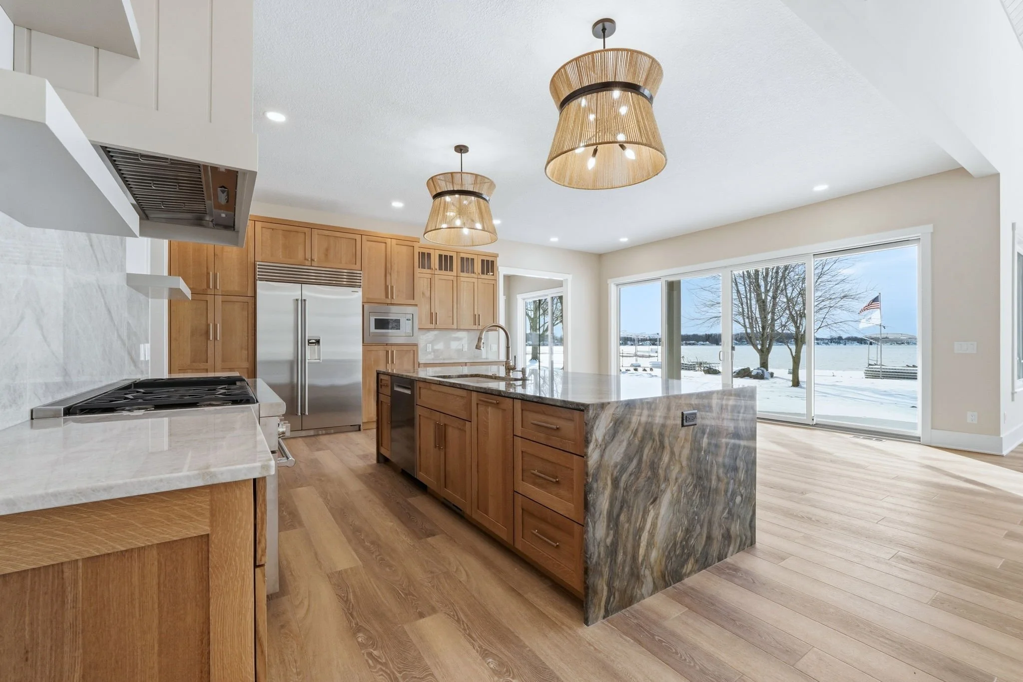 Modern kitchen with wooden cabinets, a marble island, stainless steel appliances, and large glass sliding doors leading to a snowy outdoor scene with lake view.