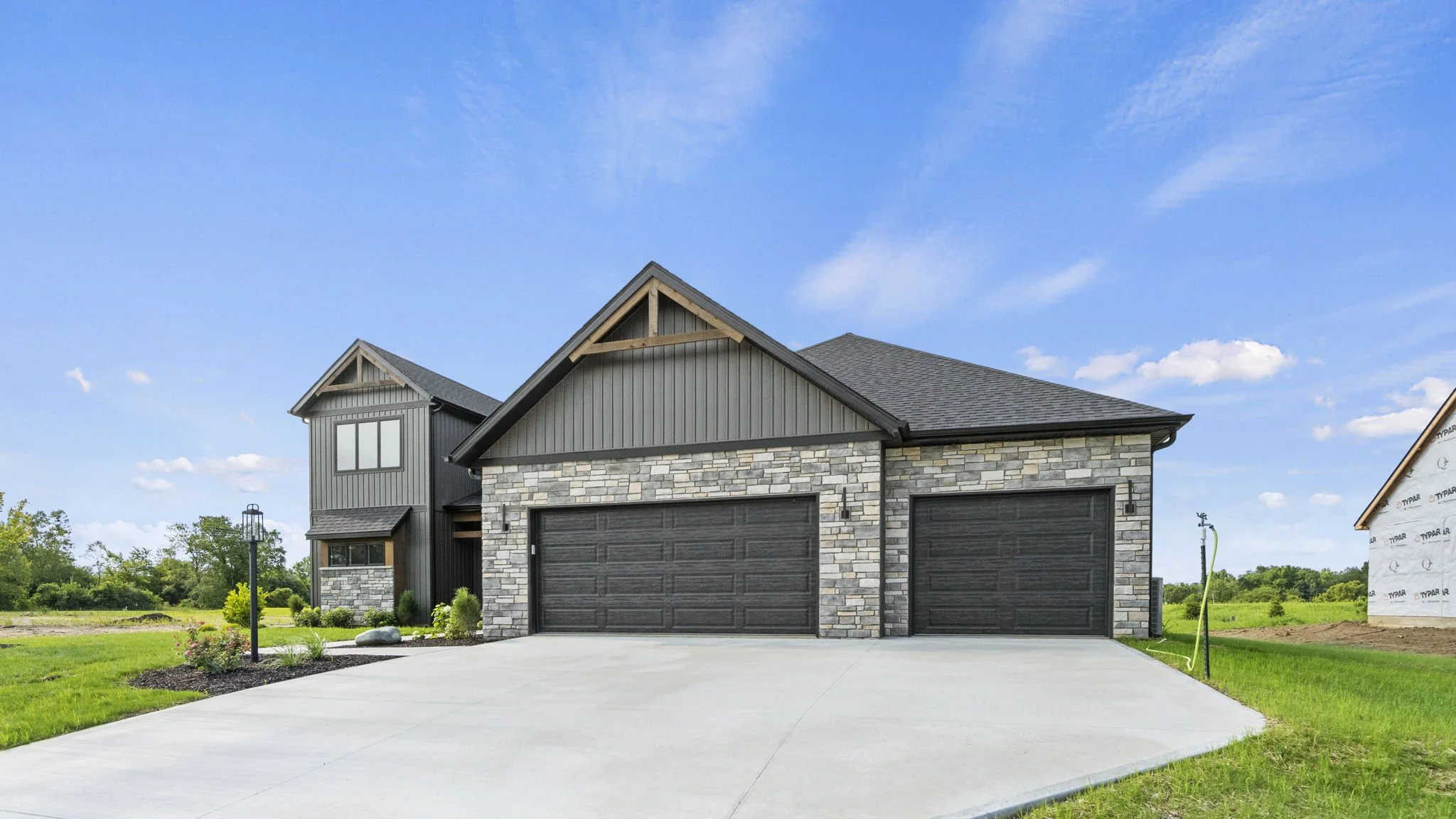 Exterior view of a modern house with a stone and gray siding facade, black garage doors, and a concrete driveway. The house is under construction with a partially finished section visible on the right. The sky is blue with some clouds, and there is a