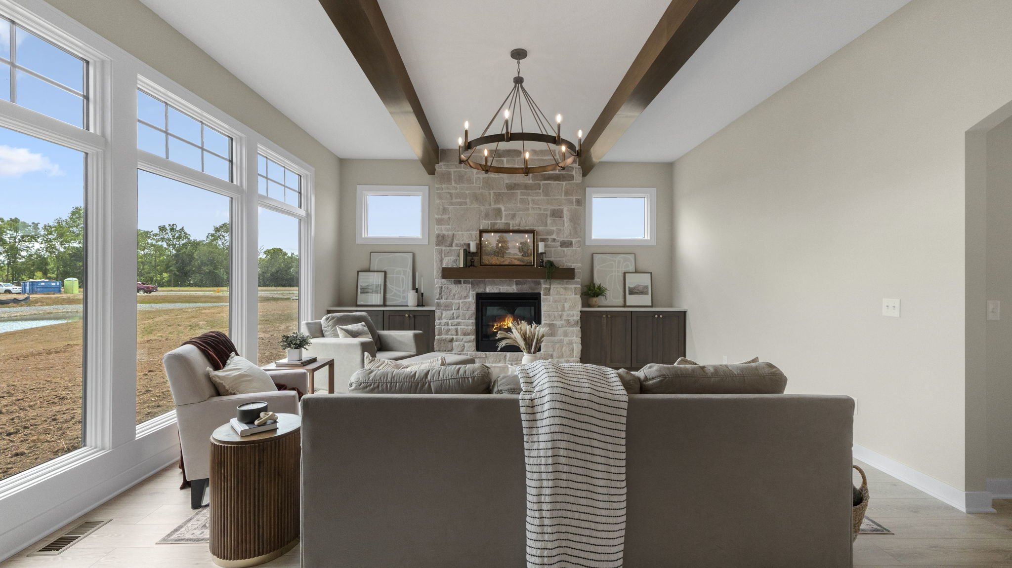 Living room with a large window showing an outdoor landscape, a stone fireplace, and beige sofas arranged around it. Overhead, a rustic chandelier hangs from wooden beams.