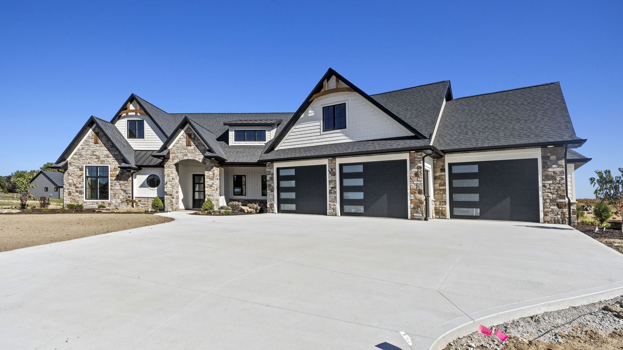 Large modern house with stone and white exterior, multiple gabled roofs, and three black garage doors, under a clear blue sky.