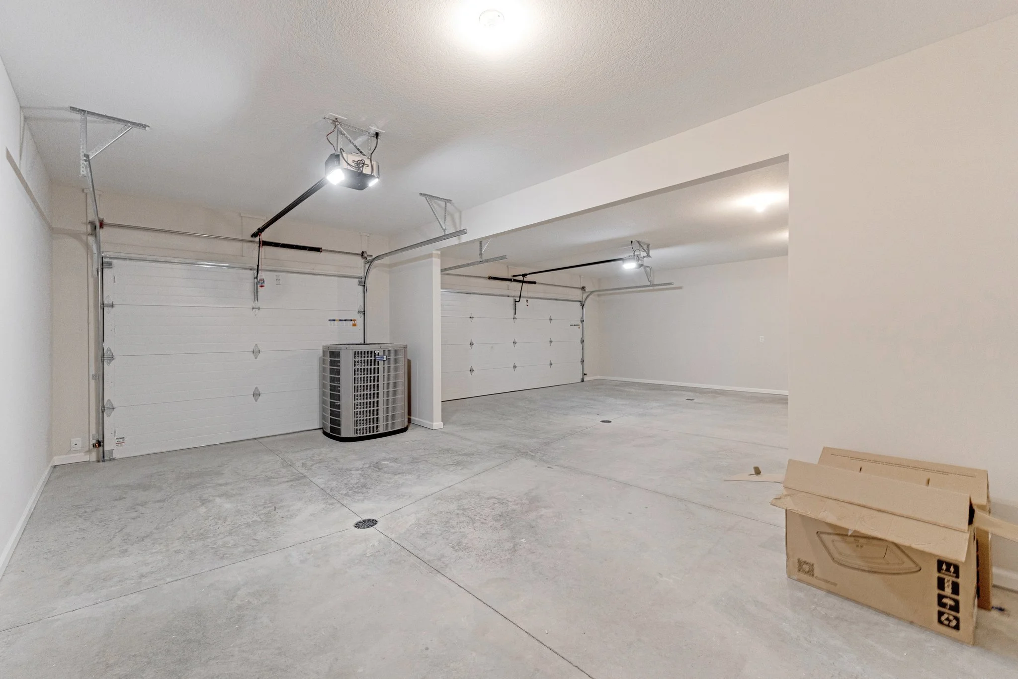 Empty garage with three white roll-up doors, a cardboard box in the corner, and a large air conditioning unit.