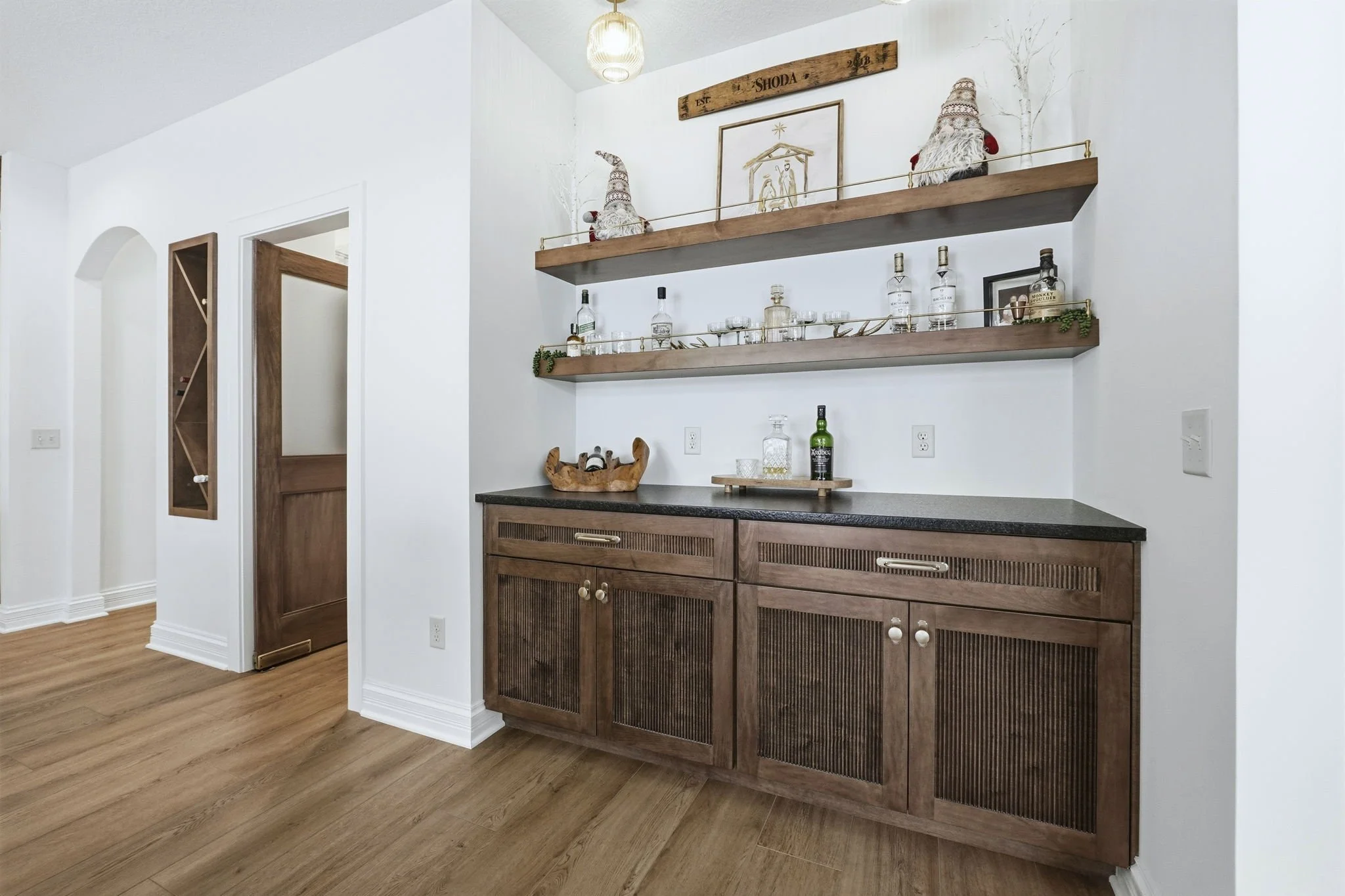 A wooden bar cabinet with a black countertop, two decorated shelves above, displaying liquor bottles, glasses, and decorative items, in a room with white walls and wooden flooring.