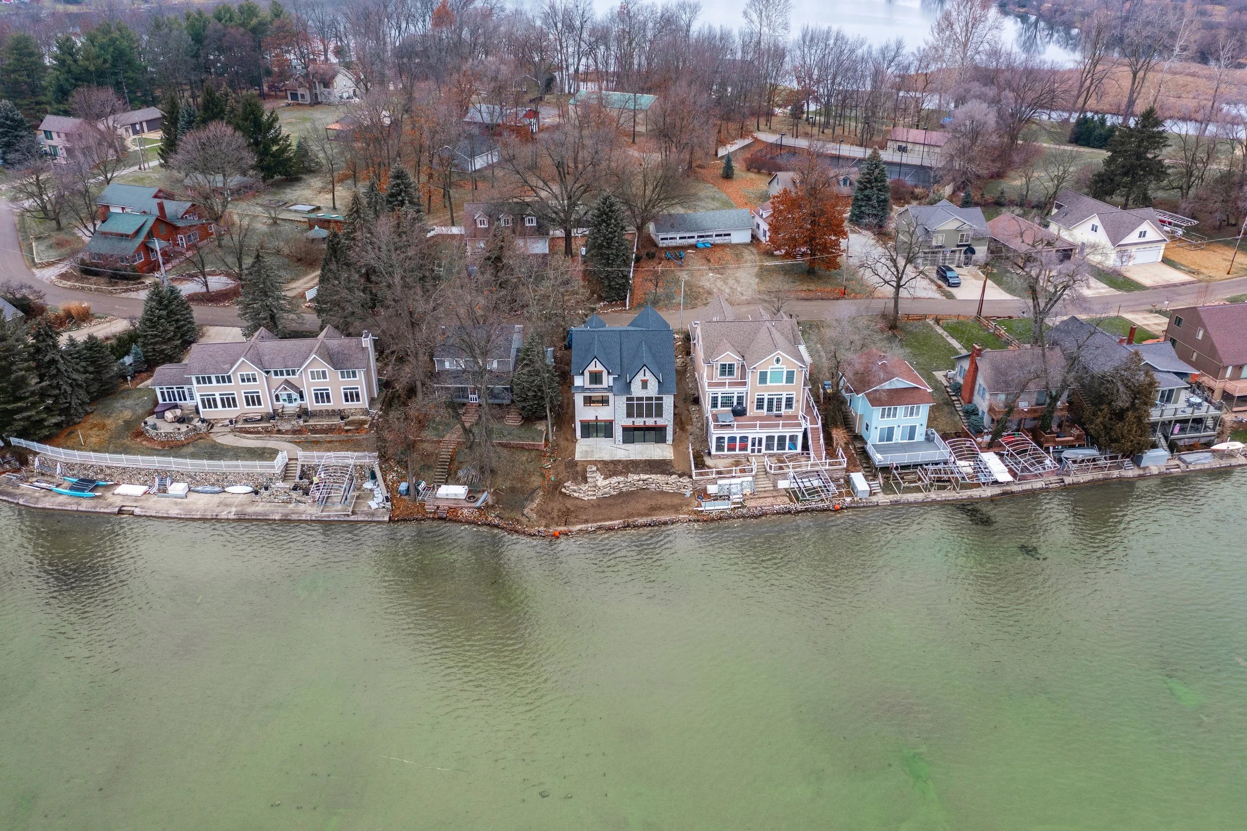 Aerial view of lakeside houses with boats, docks, and landscaped yards on a calm water shoreline with bare trees and neighboring homes in the background.