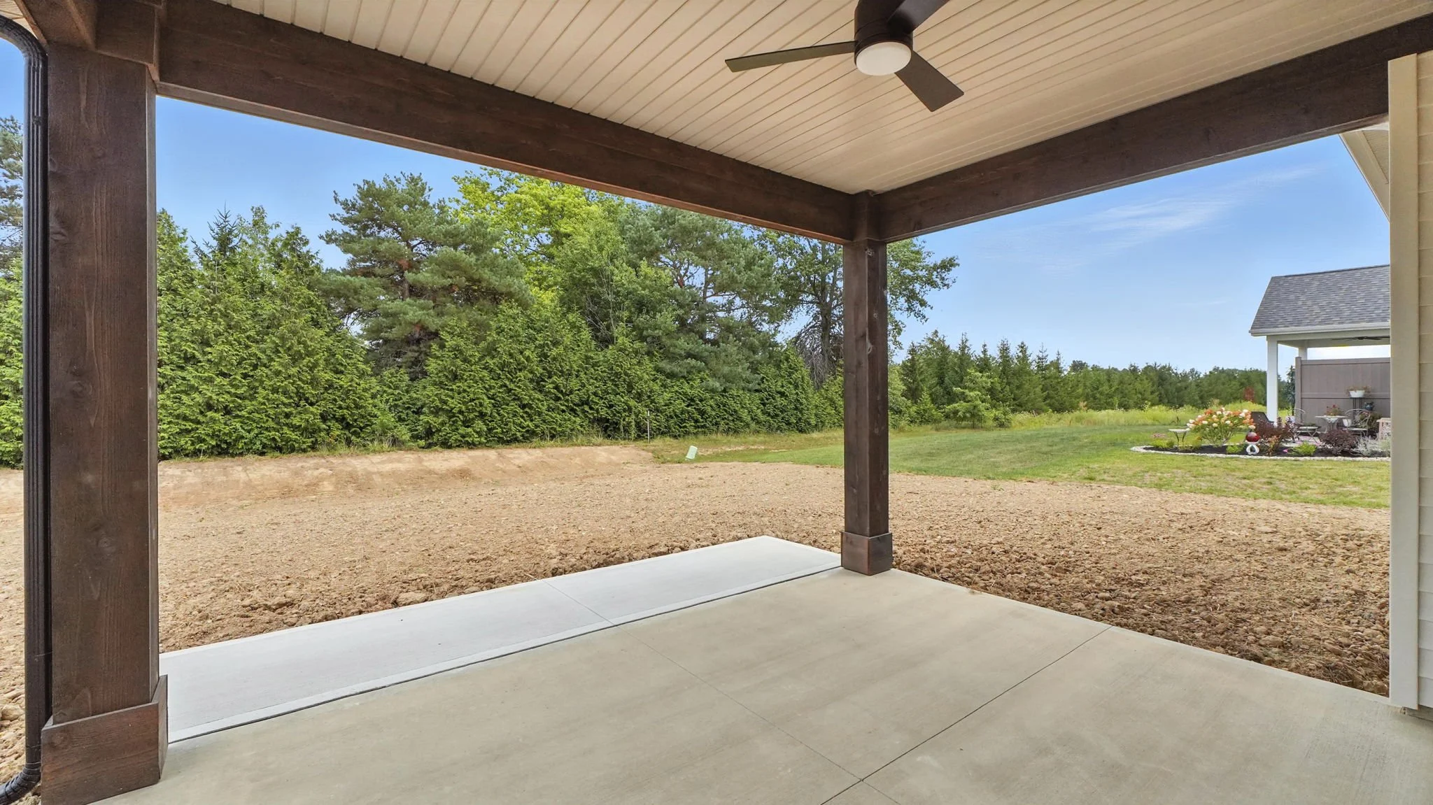 View of a backyard from a covered patio with wooden beams, concrete flooring, a dirt area, grass, trees, and a house with a garden.