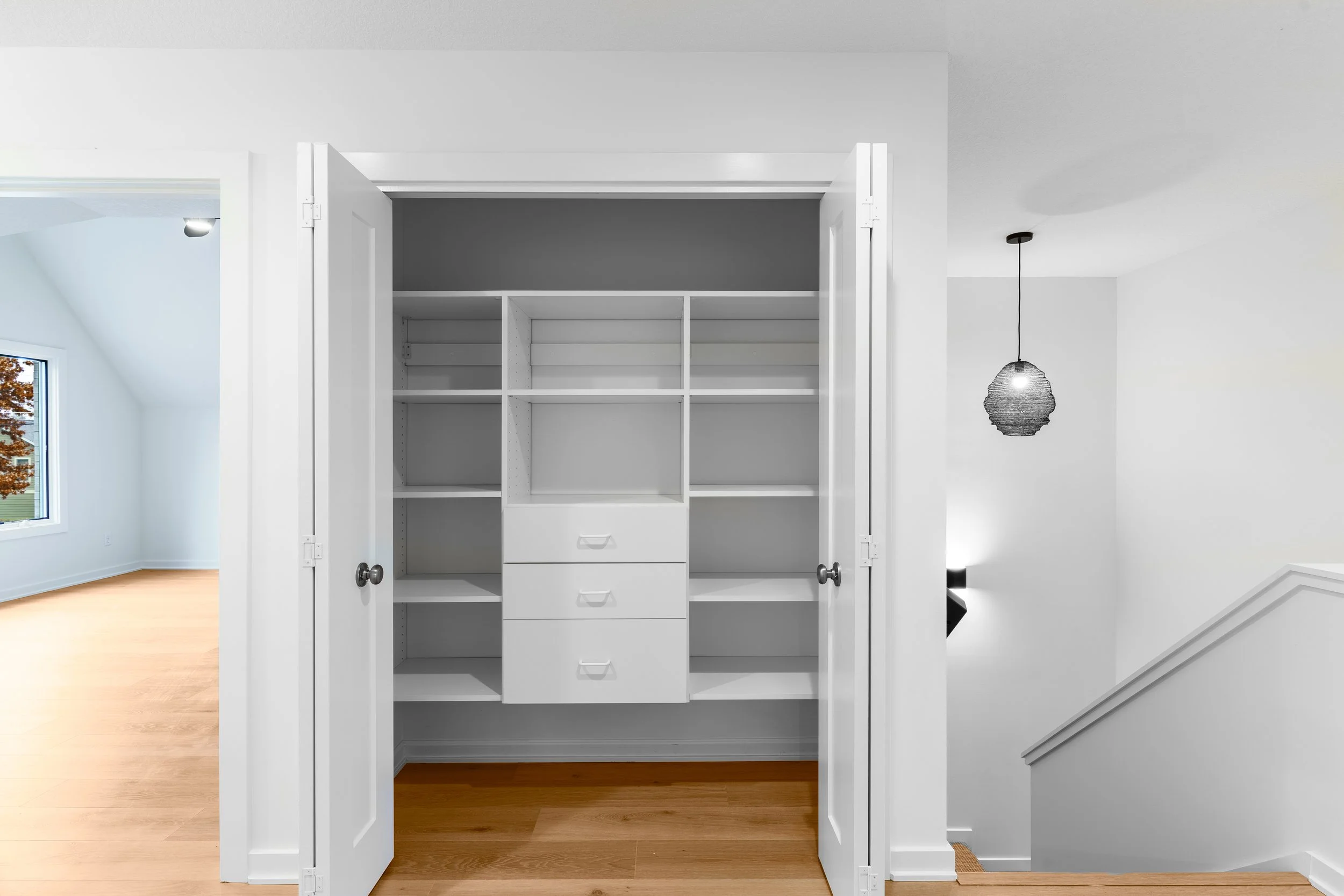 Empty white closet with open doors in a modern home, with a stairway and hallway visible in the background.