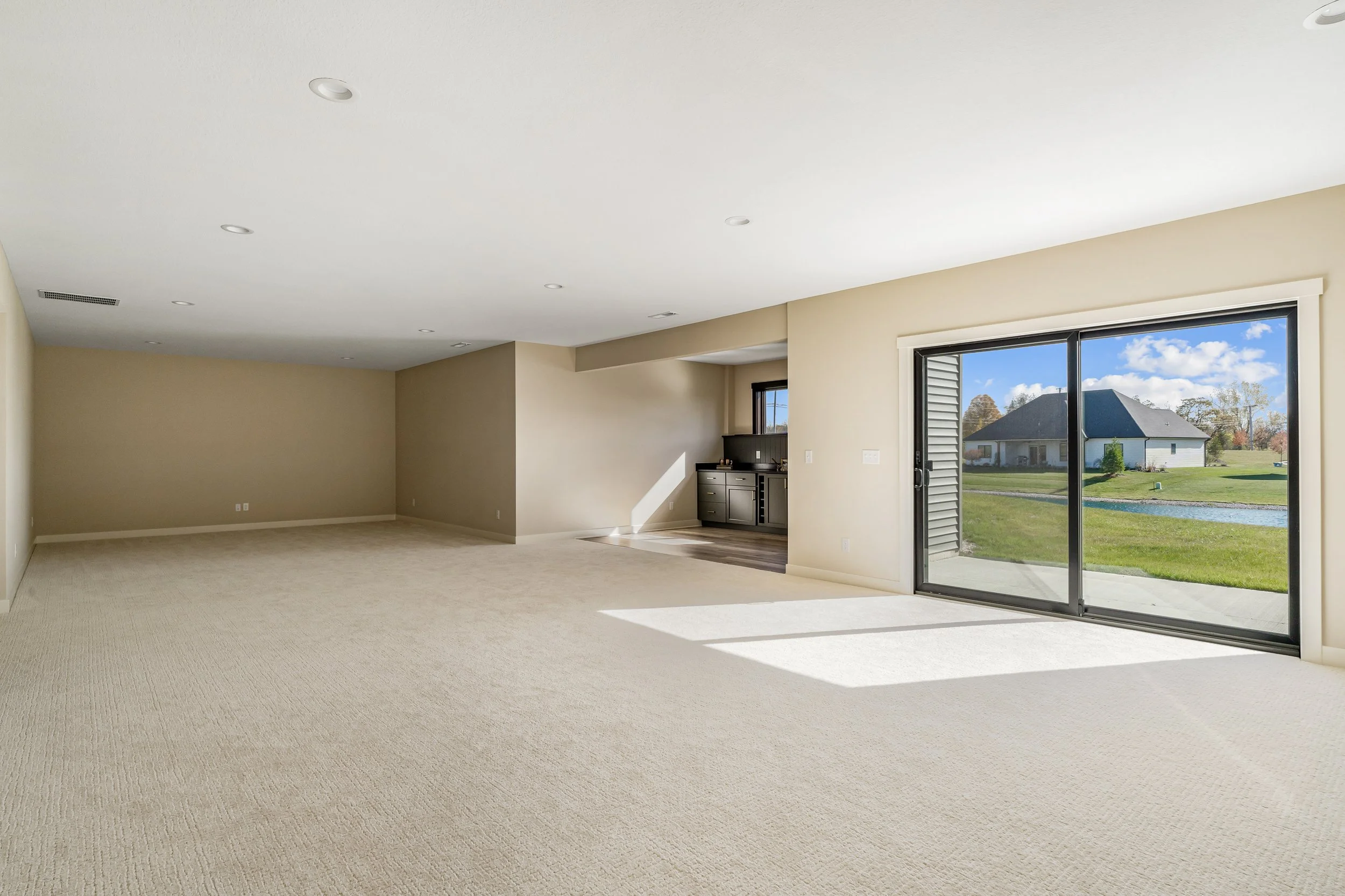Empty living room with beige walls, carpeted floor, large sliding glass door leading outside to a yard with green grass and a pond, and a small built-in black cabinet near the kitchen area.