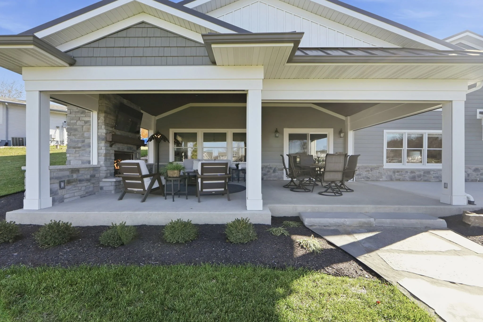 Back porch of a house with outdoor furniture, a stone fireplace, and a mounted television, with a pathway leading to stairs and a landscaped lawn.