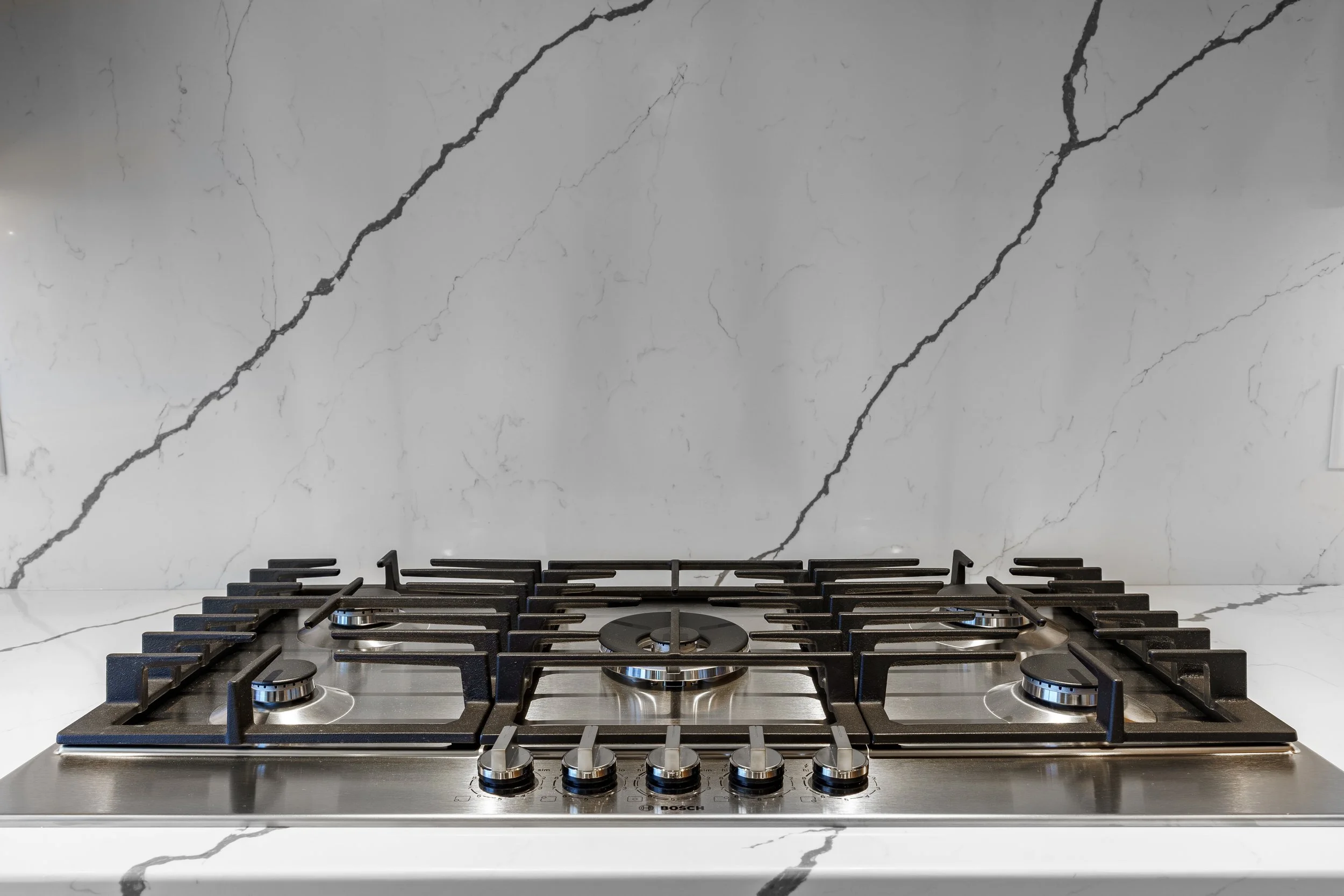 Modern stainless steel gas cooktop with five burners on a white marble countertop with gray veining.