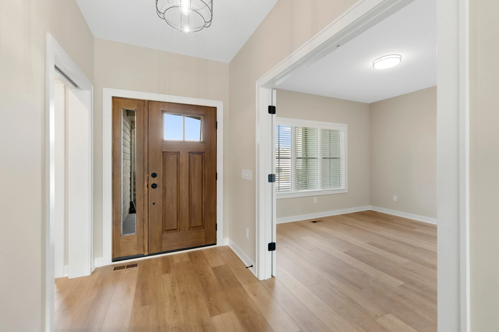 Empty living room with wooden flooring, a wooden front door with glass window, and a room with large windows and white blinds.