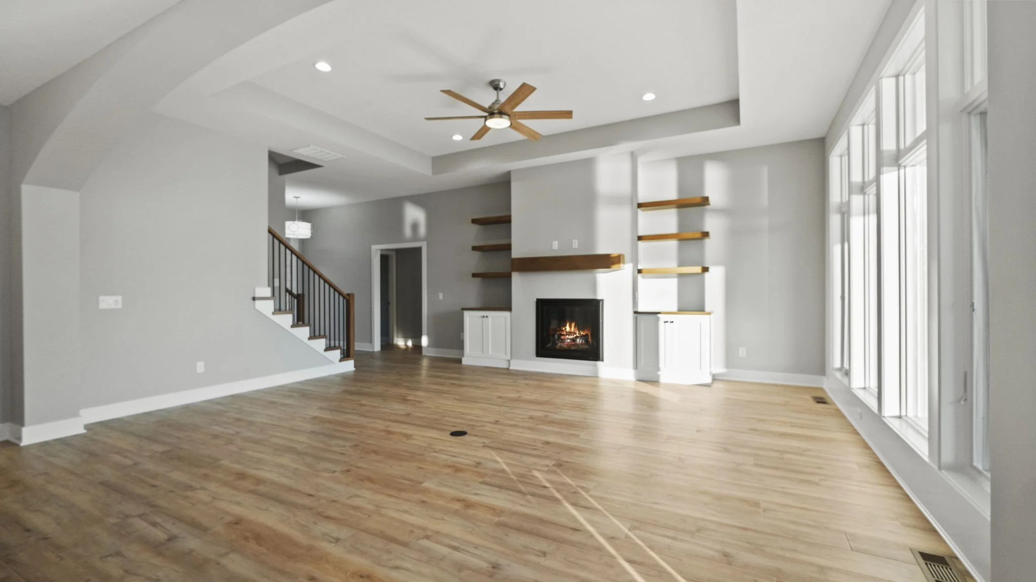 Empty living room with hardwood floors, a modern fireplace, built-in wooden shelves, large windows to the right, and a ceiling fan.