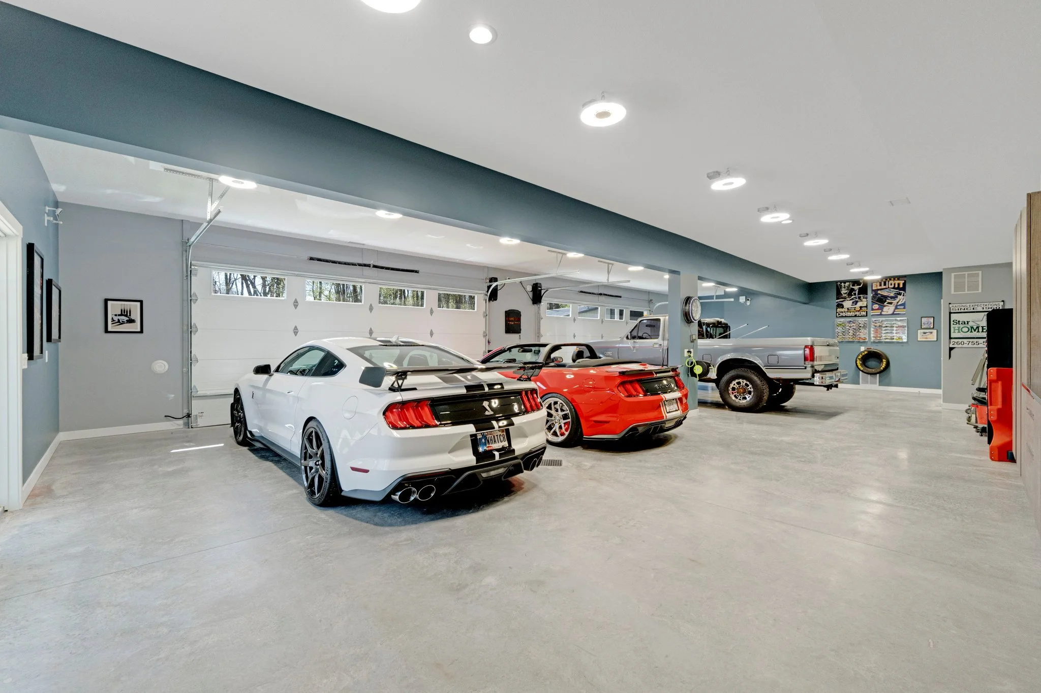 A spacious garage with three cars parked inside: a white sports car, a red sports car, and a gray pickup truck. The garage has a white garage door, gray walls, and ceiling lights.