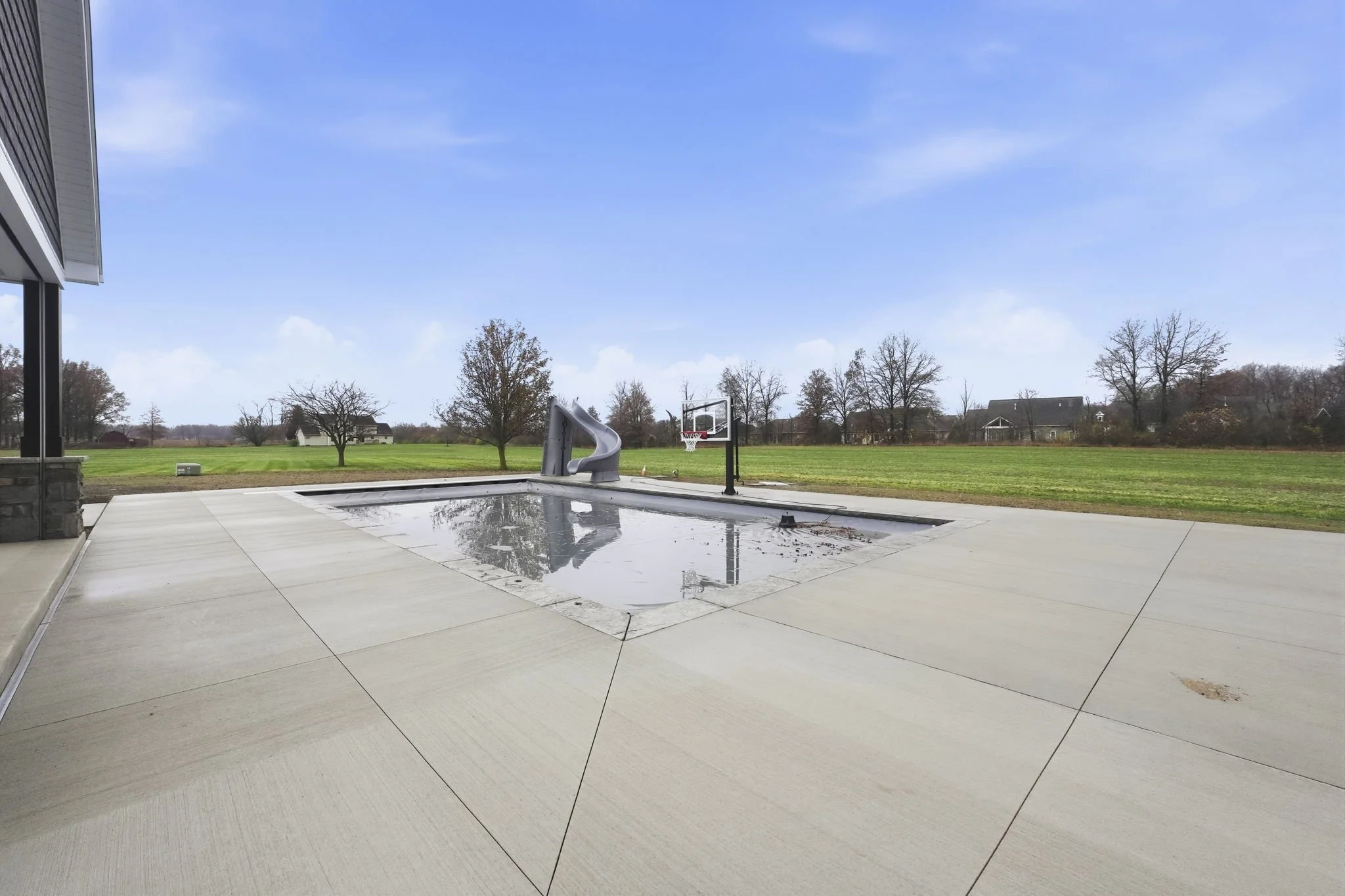 Empty backyard with swimming pool covered in water, basketball hoop, and a playground slide, with trees and houses in the background under a blue sky.