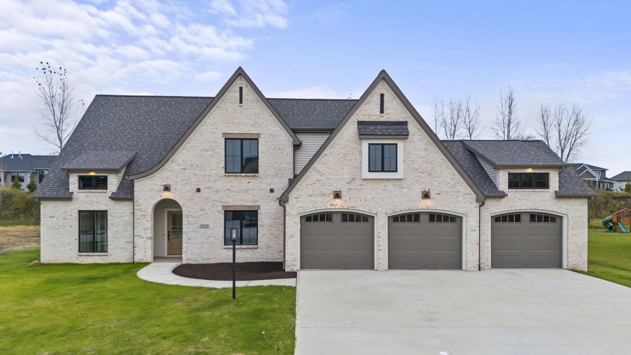 Modern white brick house with three garages, black framed windows, and a landscaped front yard under a blue sky.
