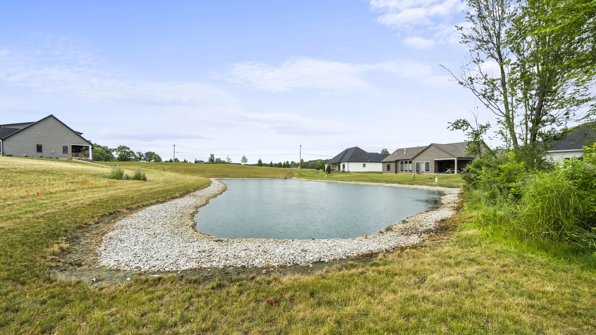 Residential neighborhood with houses and a pond in the foreground, grassy landscape, trees, and a blue sky with some clouds.