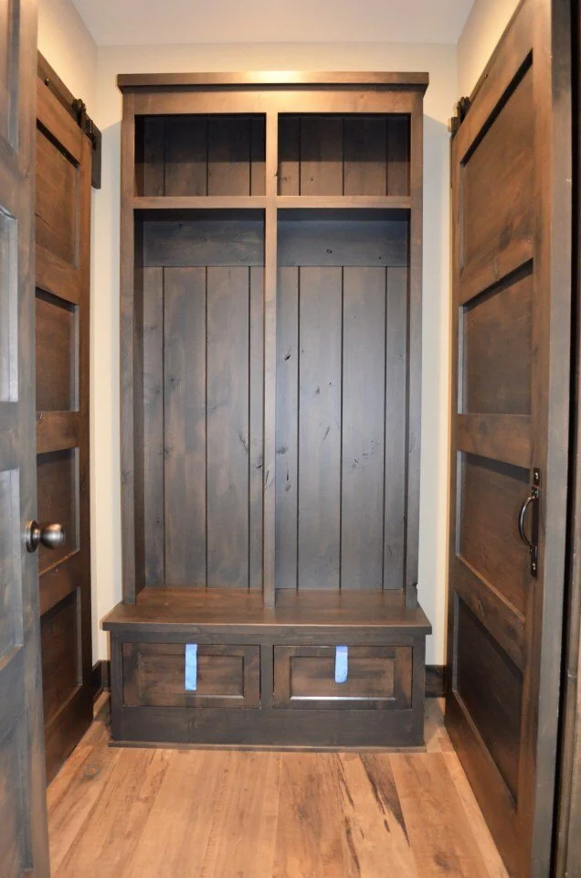 A wooden mudroom bench with hooks and storage, flanked by two framed wooden doors, in a hallway with hardwood flooring.