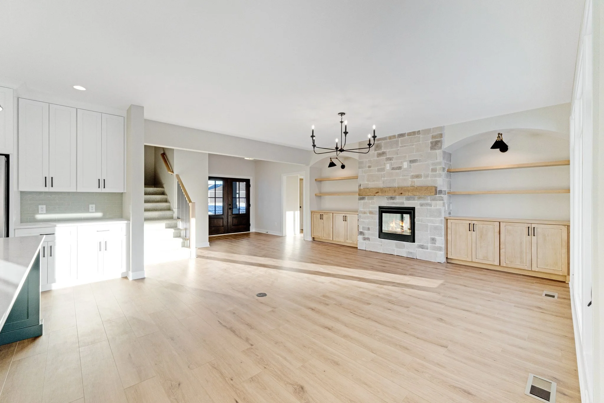 Bright, open living room with light wood flooring, a stone fireplace, built-in wooden cabinets and shelves, a black chandelier, and large glass doors leading outside.