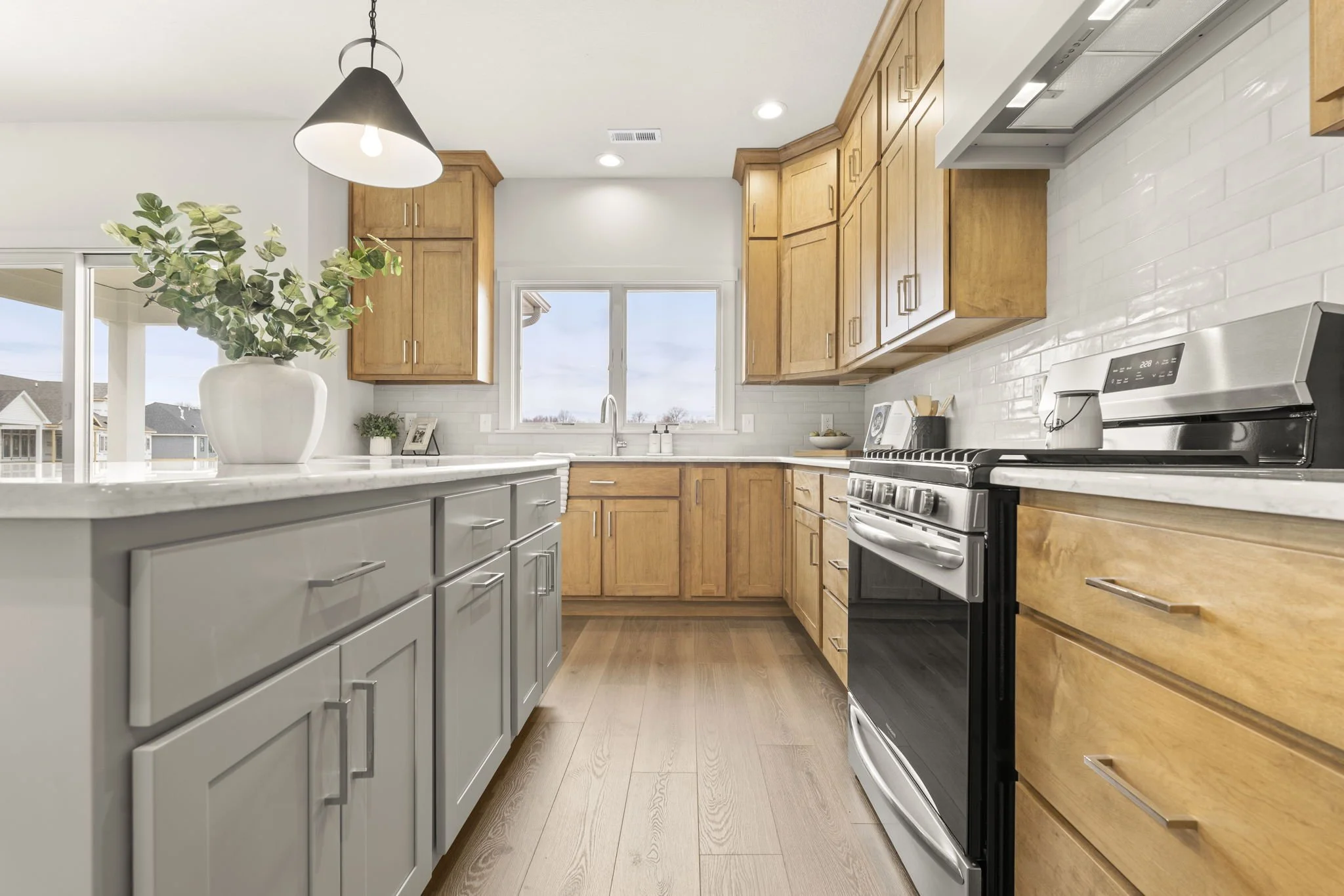 Modern kitchen with white and light brown cabinets, a white countertop, a black stove, a window above the sink, a white vase with greenery on the counter, and hardwood flooring.