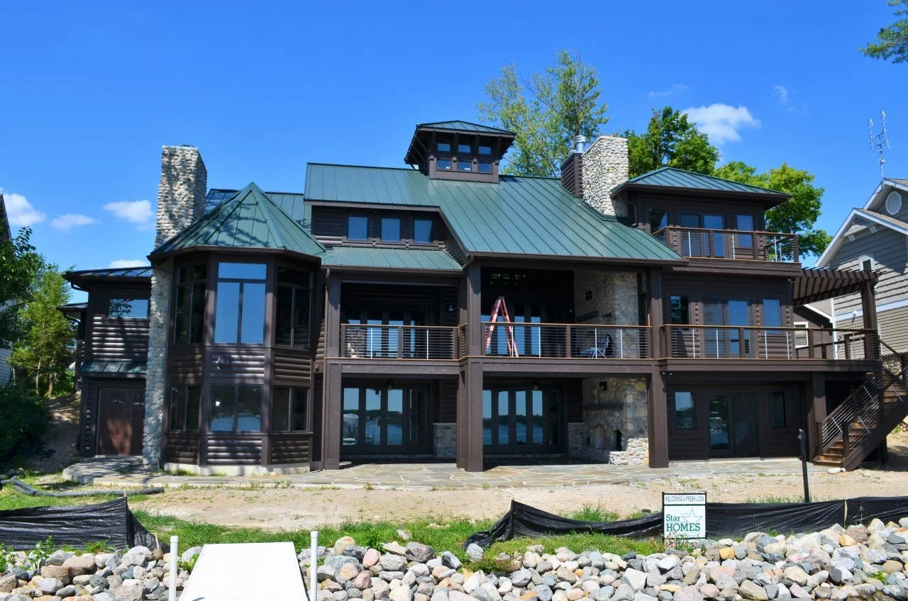 Large modern house with stone and dark wood exterior, multiple levels, and large windows, situated on a clear day with a bright blue sky and green trees in the background.