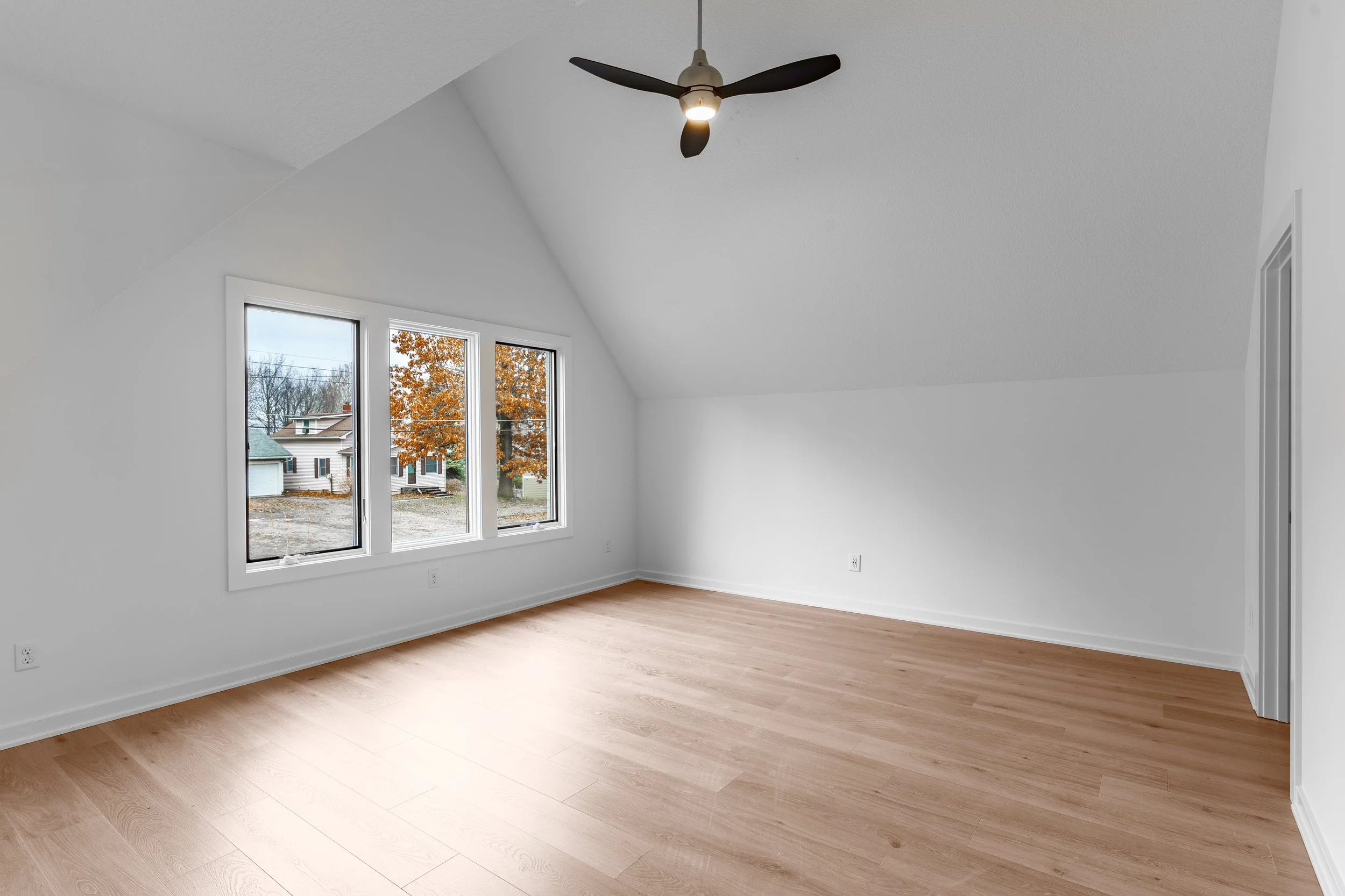 Empty bedroom with white walls, light wooden floors, three large windows, a ceiling fan with black blades and a light, and a partly closed door.