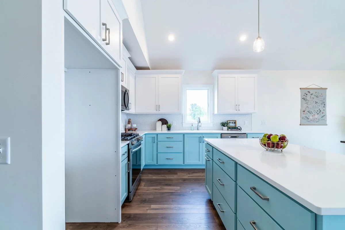 Kitchen with white and light blue cabinets, white countertop, window, apples in a wire basket, and a hanging pendant light